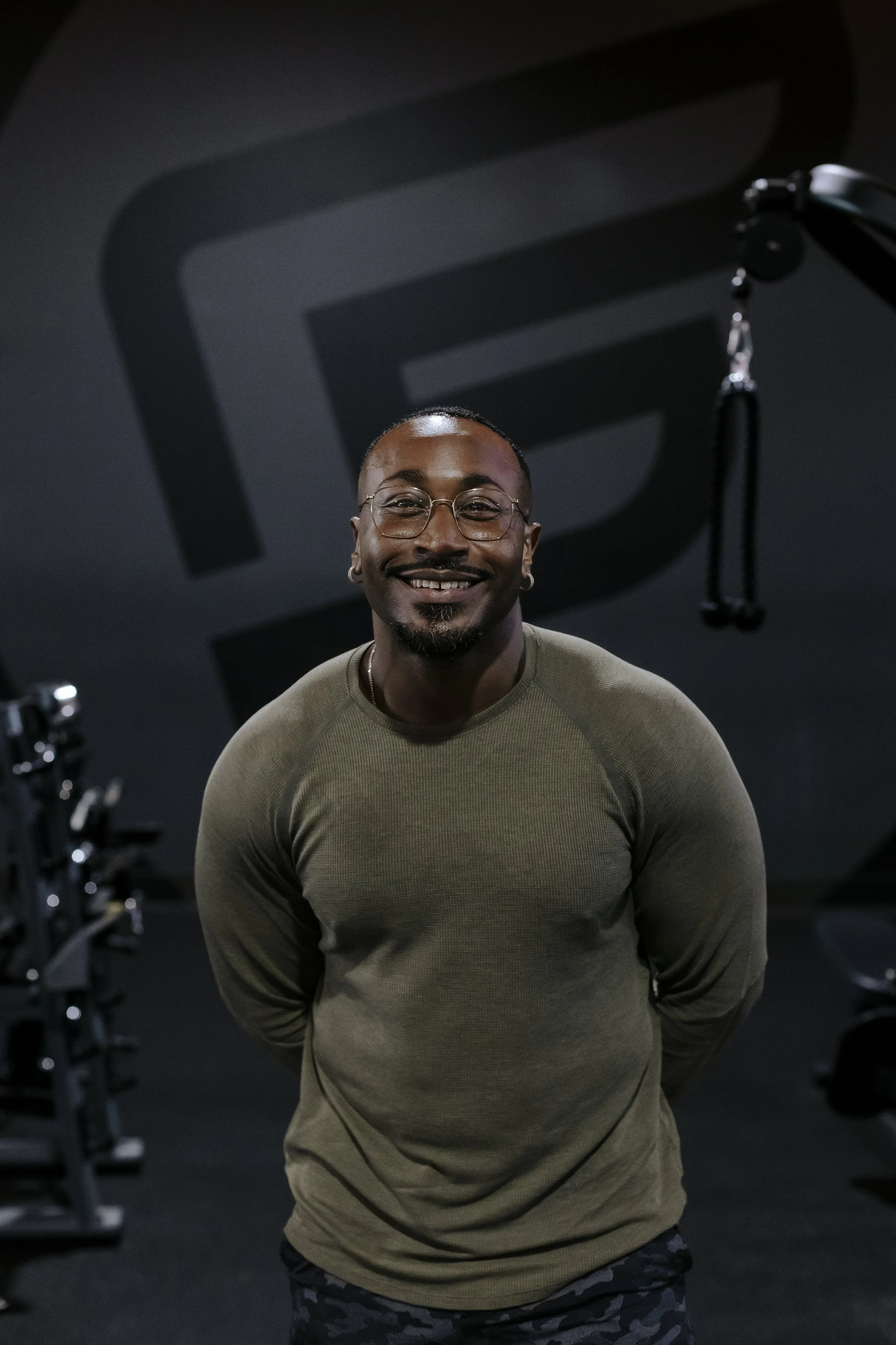 A smiling man with glasses, earrings, and a beard standing in a gym with fitness equipment and a large logo on the wall behind him.