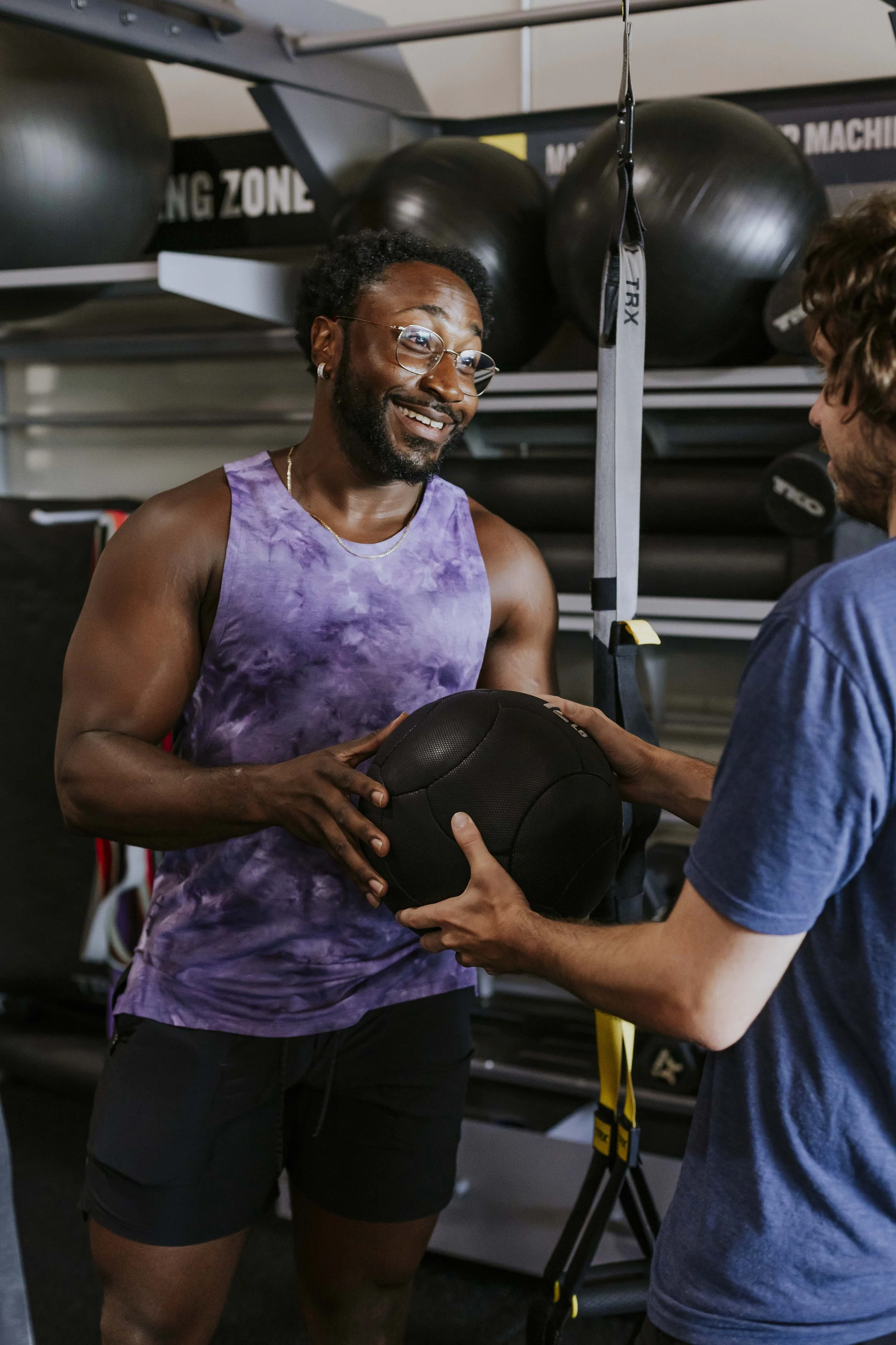 A man in a purple tie-dye tank top and black shorts is holding a medicine ball with both hands. Another person in a blue shirt is handing the ball to him in a gym, with large black exercise balls on shelves in the background.