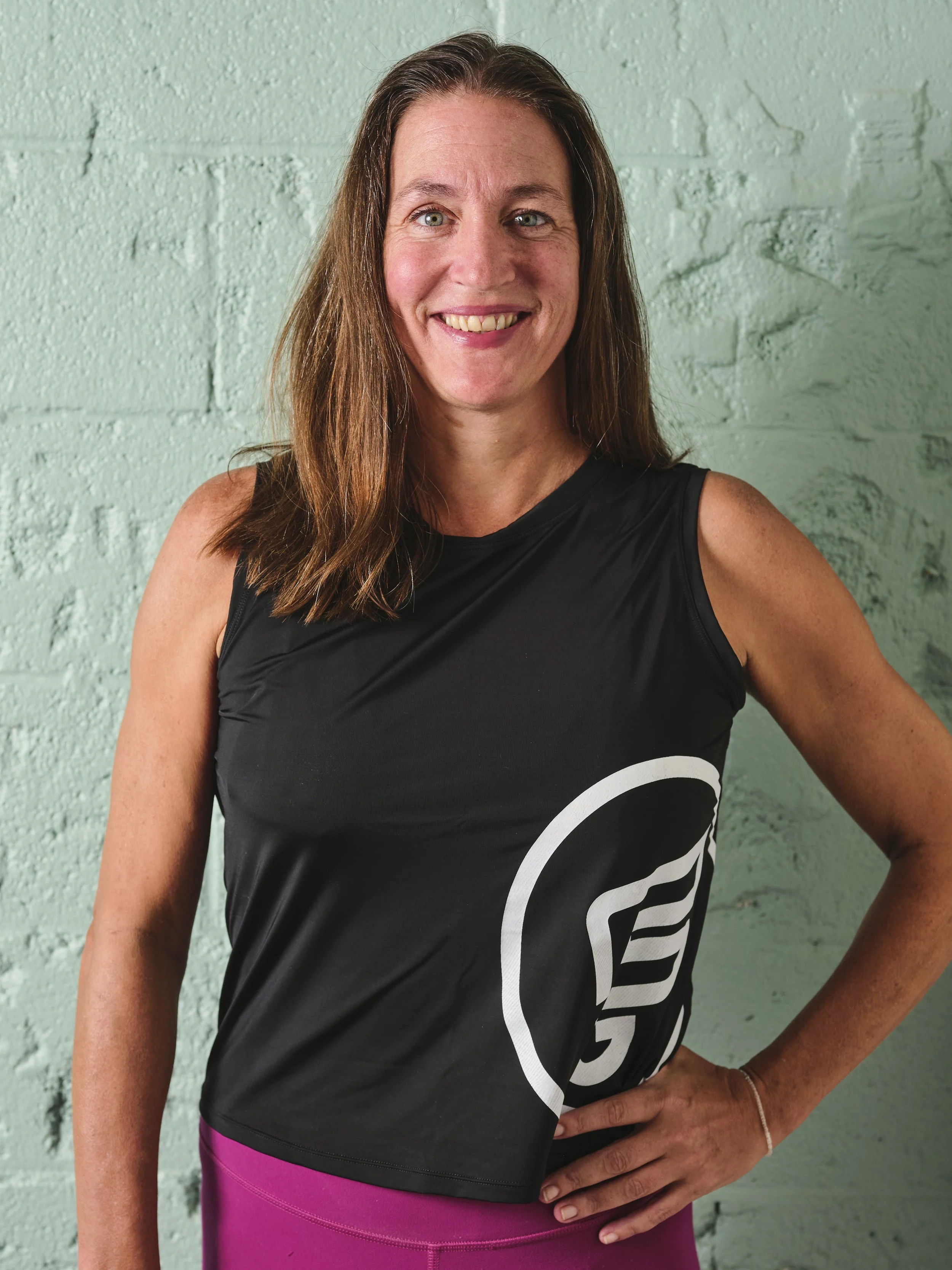 A woman with long brown hair smiling, wearing a black tank top and pink workout pants, standing against a light green textured wall.