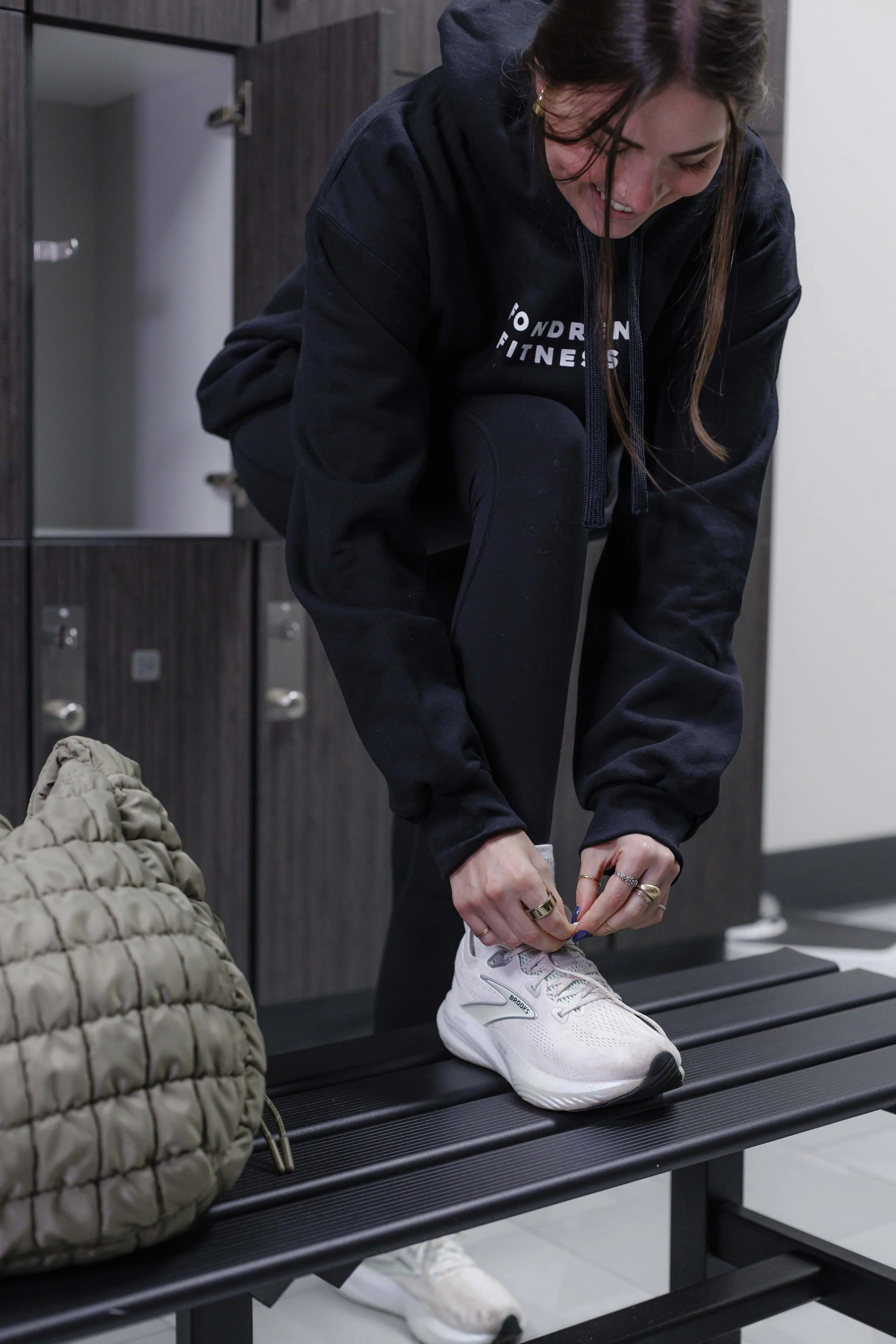 A woman wearing a black hoodie with the words 'WONDRN FITNESS' is tying her white athletic shoe on a black bench in a locker room. A beige quilted bag is on the bench, and dark wooden lockers are in the background.