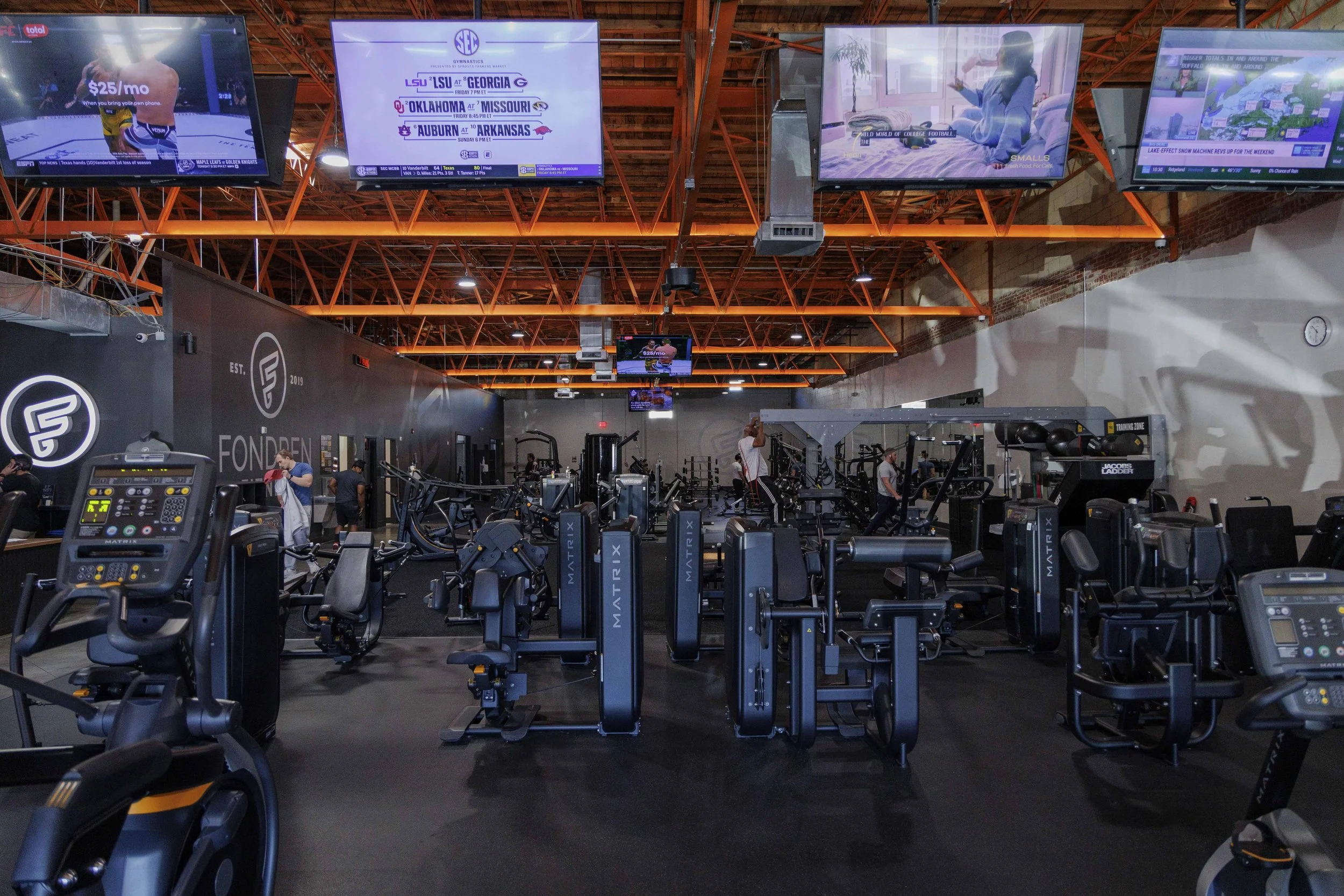 Inside a gym with treadmills and weight machines, multiple large screens hanging from the ceiling showing various advertisements and information, some workout equipment, and a few people using the machines.