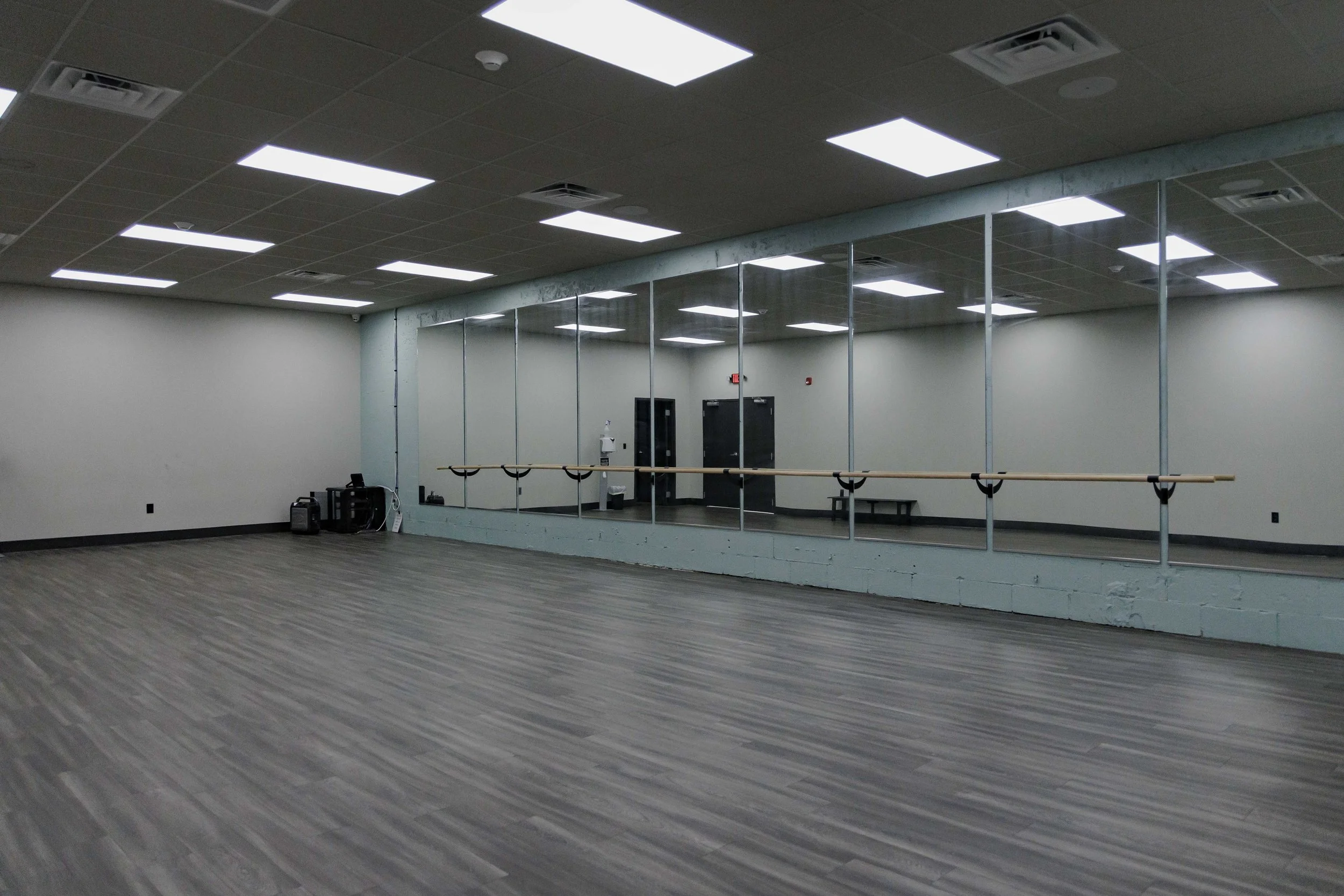 Empty dance or exercise studio with wood flooring, large mirror wall, ballet barres, and fluorescent ceiling lights.