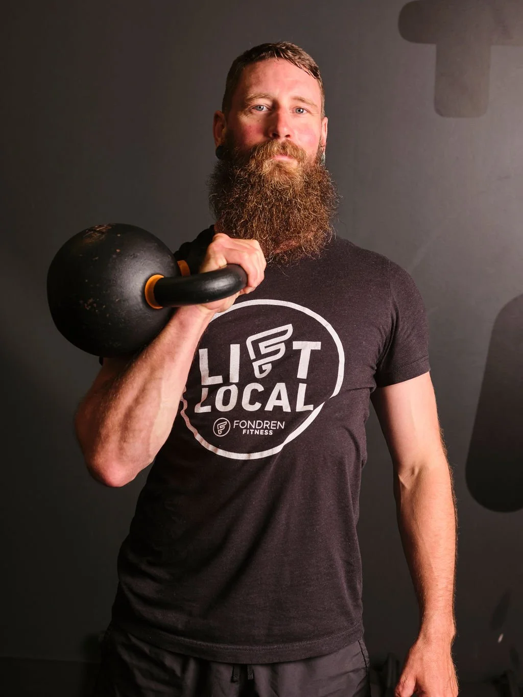 A bearded man in a black t-shirt with 'Lift Local' printed on it, holding a kettlebell over his shoulder, standing against a dark background.