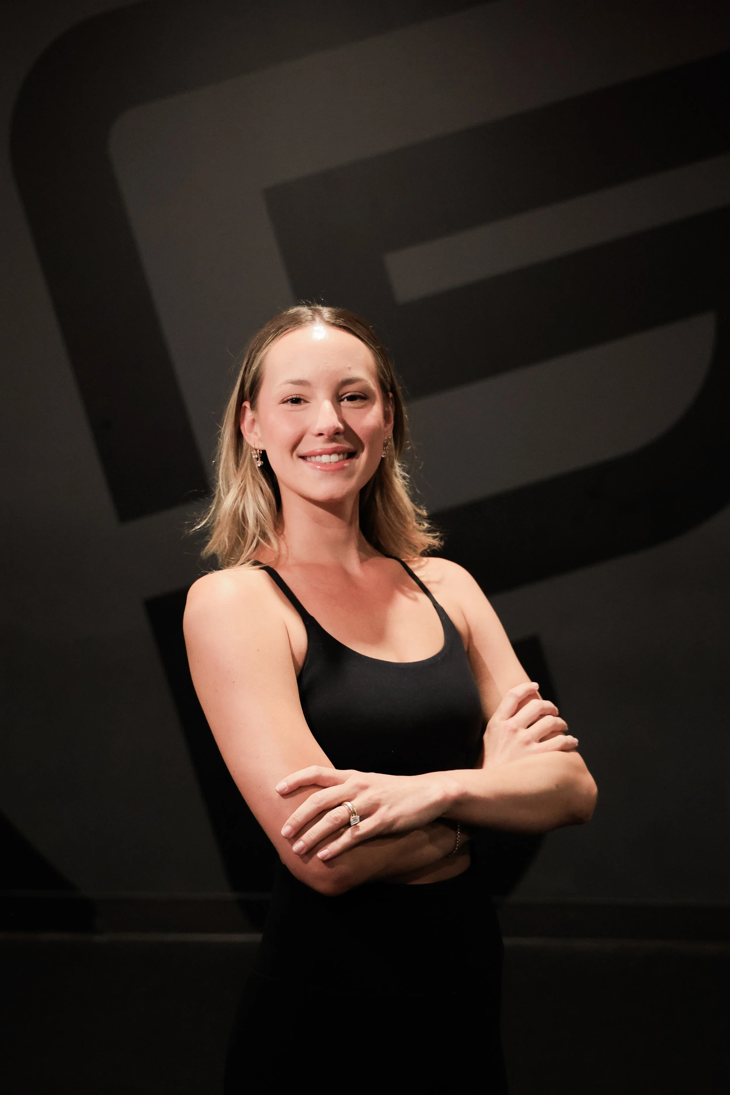 A young woman with blonde hair smiling with arms crossed in a gym setting, wearing a black sports top and jewelry, with gym equipment and TV screens visible in the background.