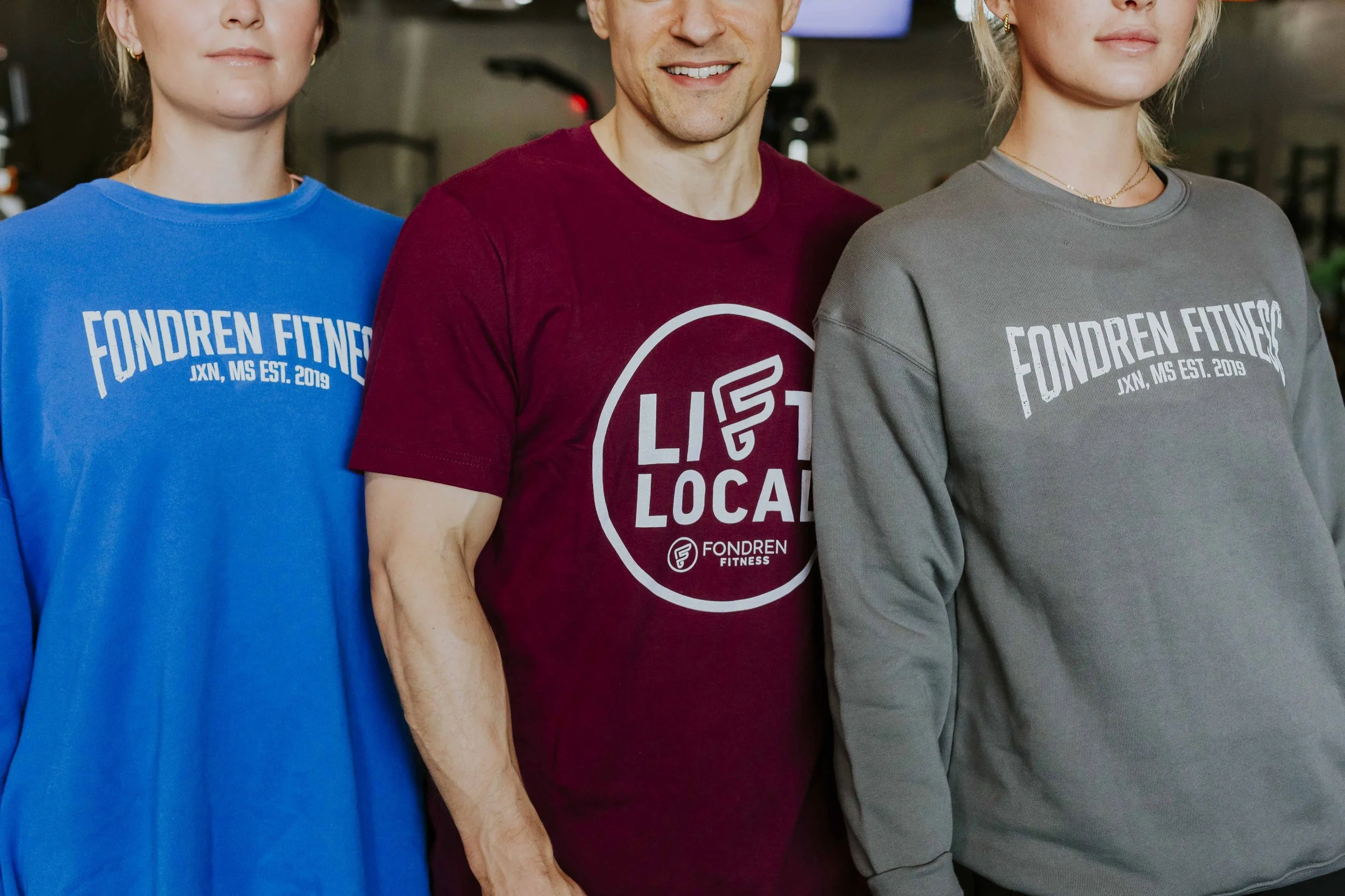 Three people standing together wearing athletic sweatshirts with fitness logos, in a gym setting.