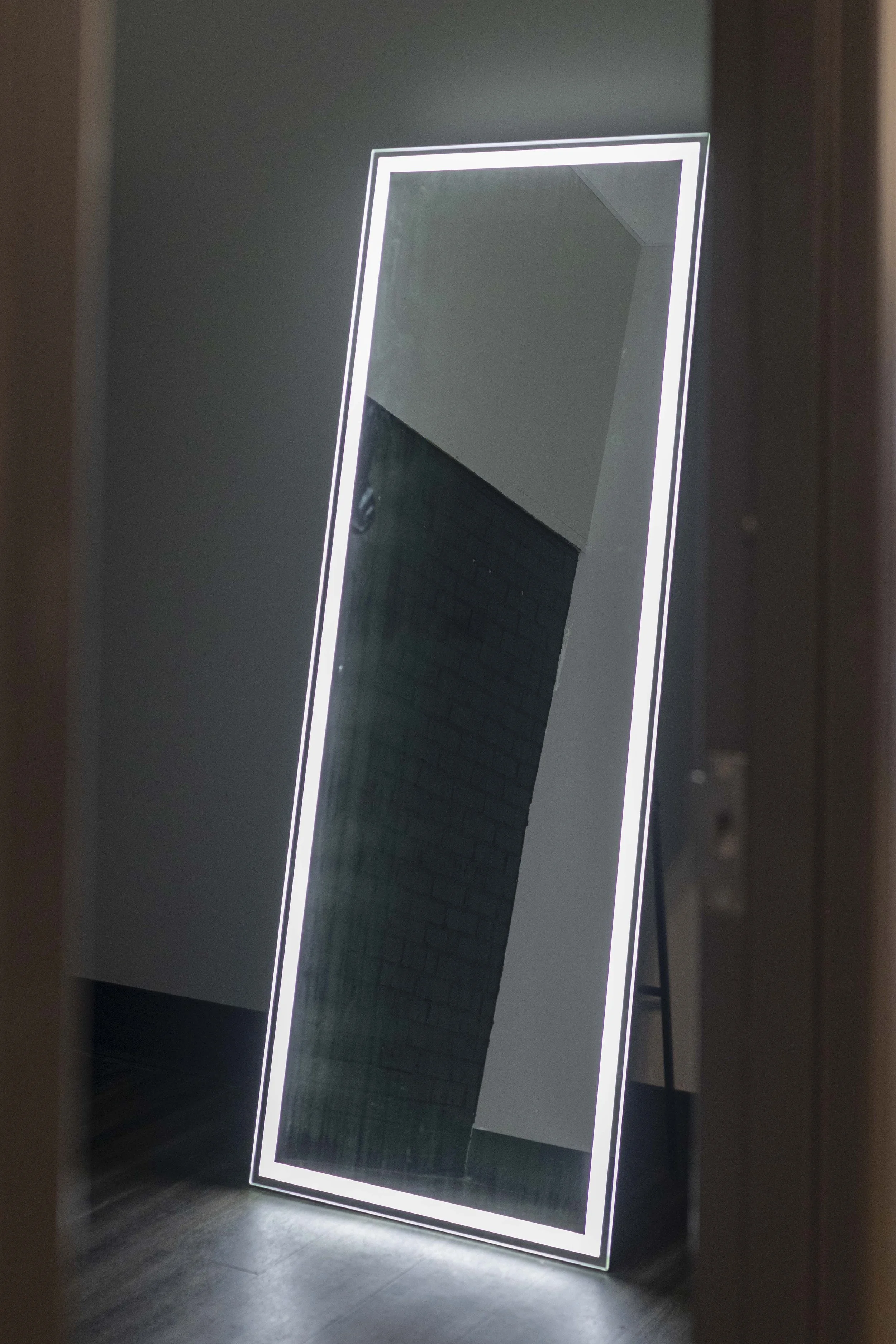 A tall, rectangular mirror with built-in LED lighting along the edges, leaning against a wall in a dark room, reflecting part of a ceiling and a brick wall.