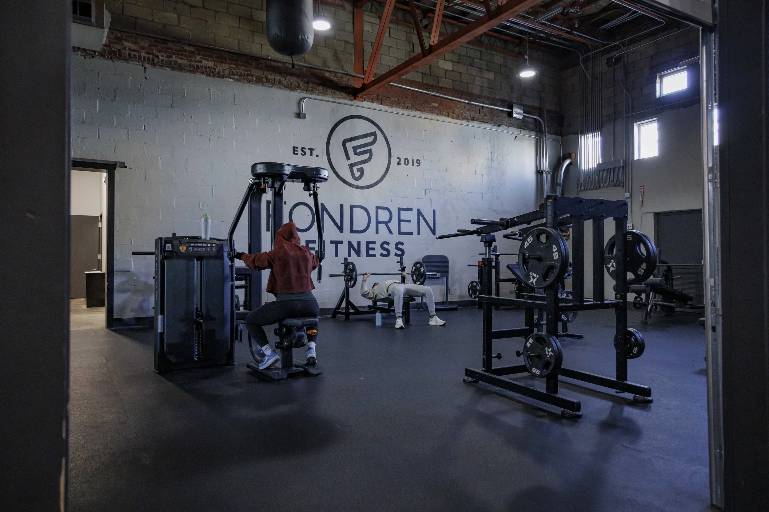 Interior view of a gym with weightlifting and workout equipment, a person in a red hoodie and gray sweatpants lying on a bench, and a person in a gray hoodie and black pants using a machine, with a large logo that reads 'LONDREN FITNESS' on the white
