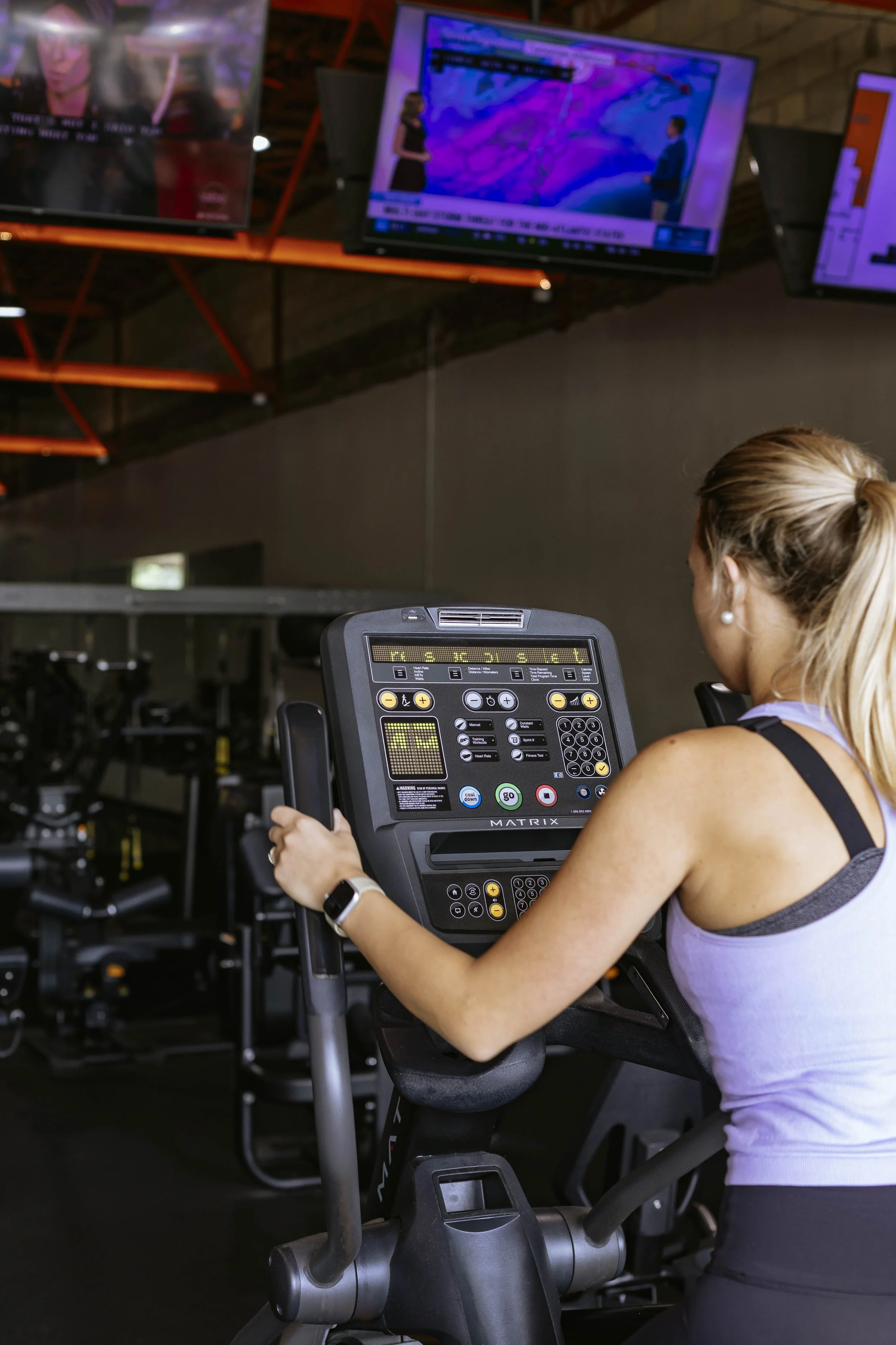 A woman using a stationary exercise bike in a gym, with multiple TV screens mounted on the wall displaying various programs and news.
