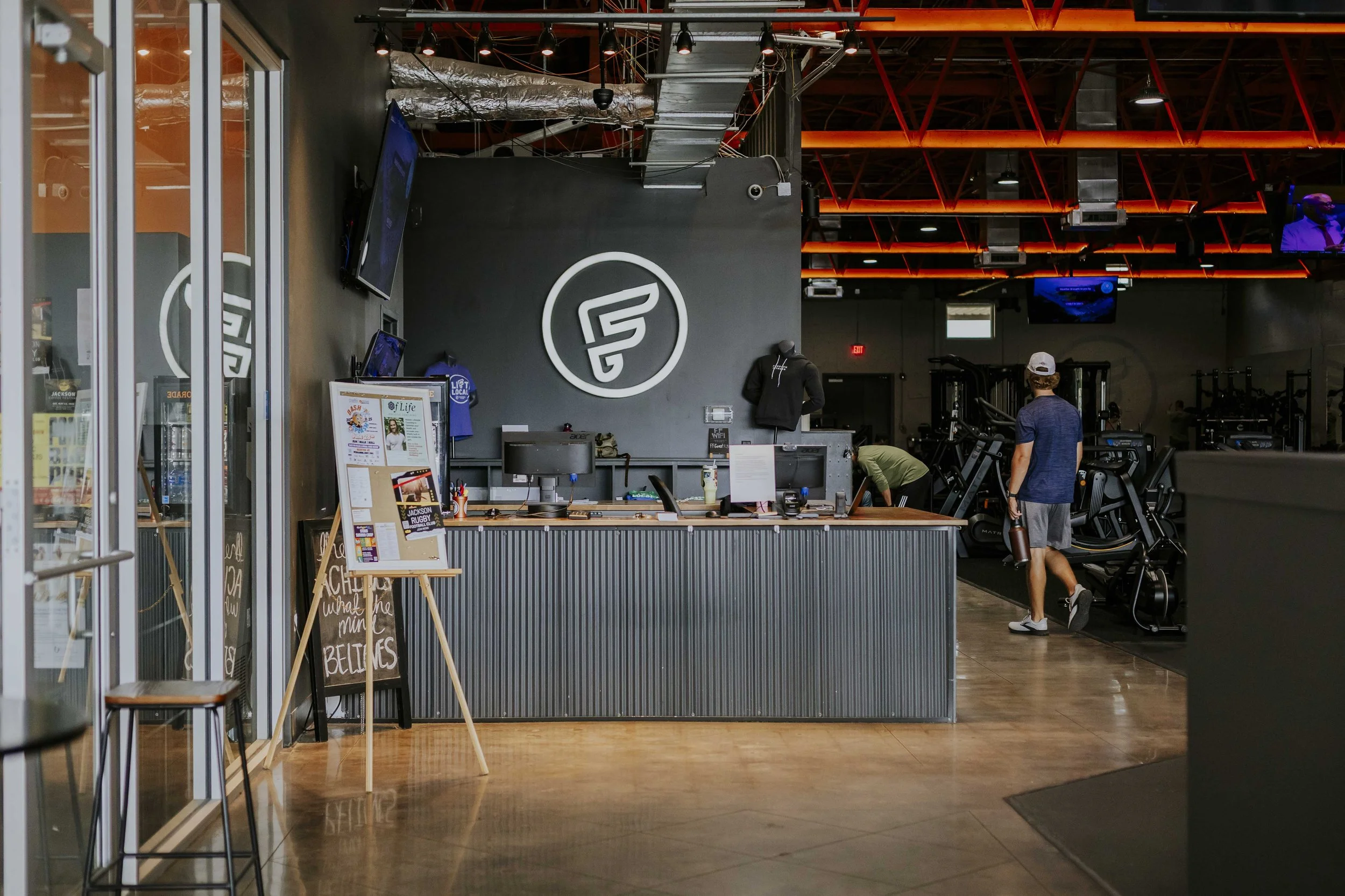 Interior of a fitness gym with a reception counter, gym equipment, and people working out. The gym has a modern industrial design with exposed ceiling pipes and orange lighting.
