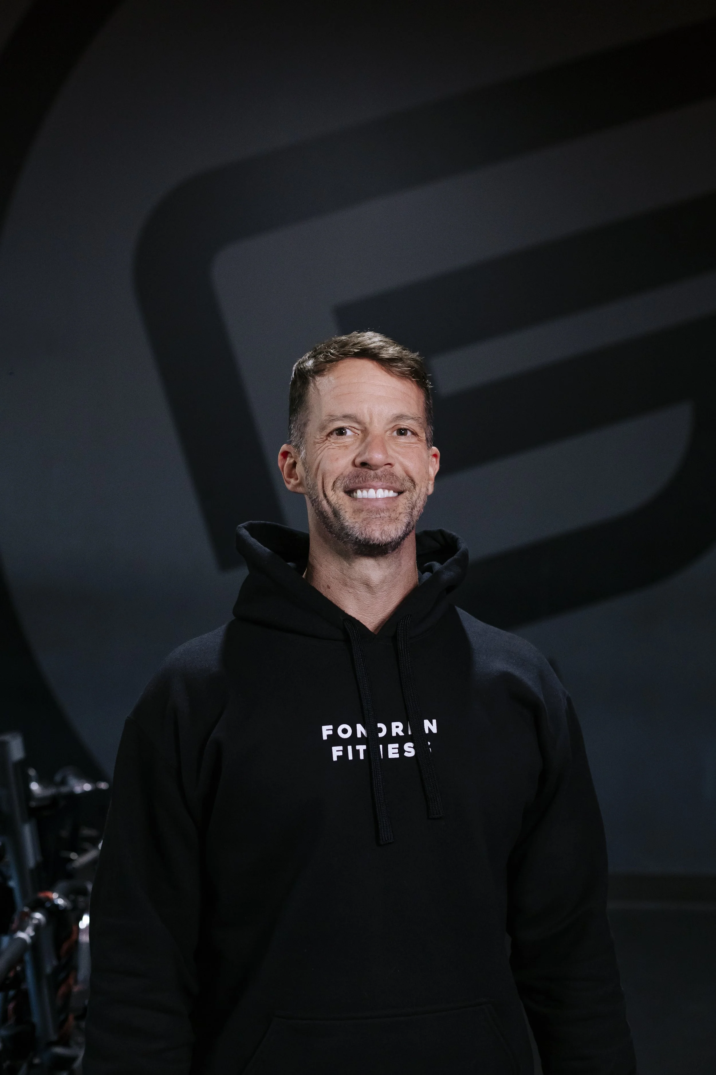 A smiling man with short brown hair and a beard, wearing a black hoodie with white text, standing in a gym with dark lighting and exercise equipment in the background.