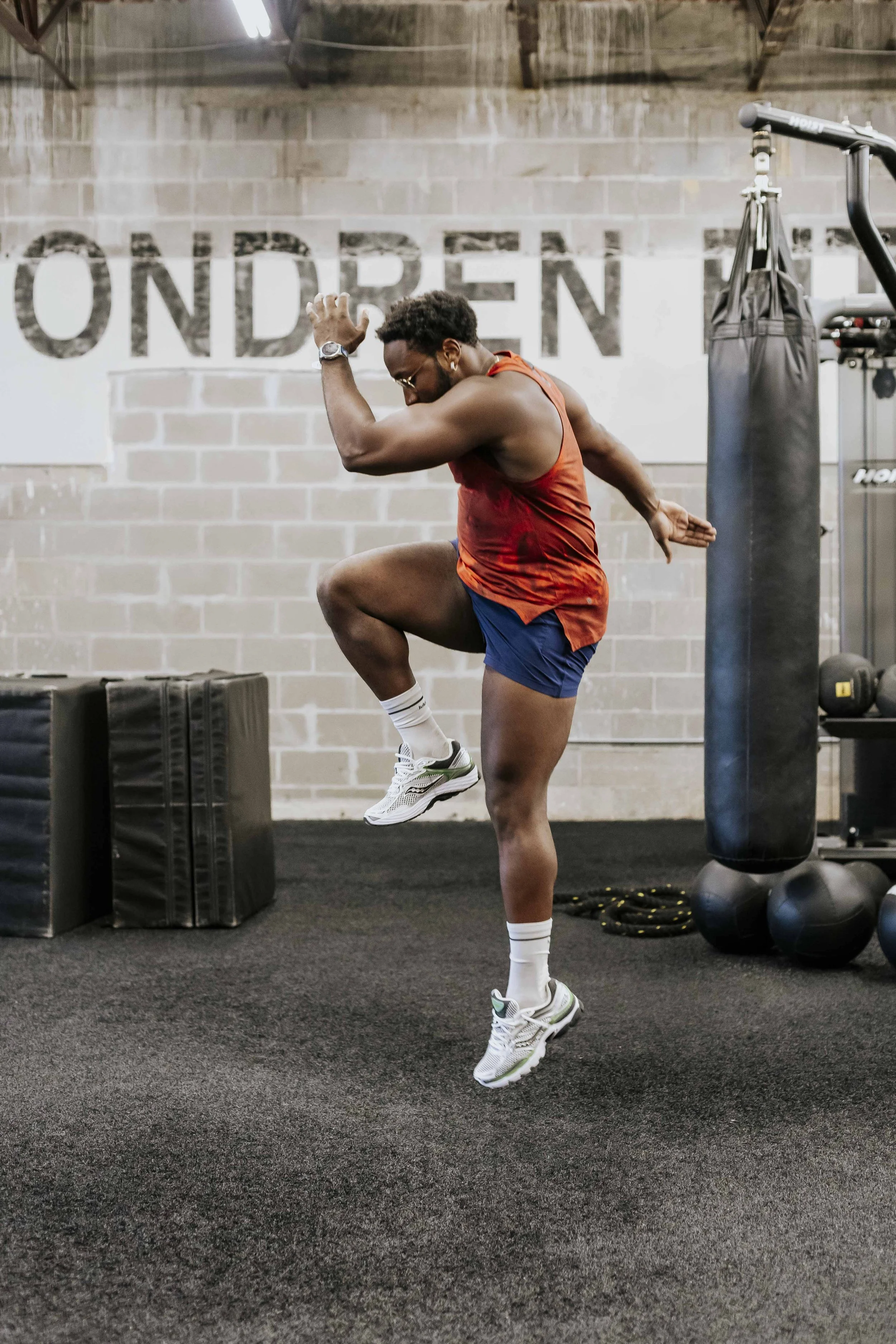 A man exercises in a gym, balancing on one leg with his chicken raised, wearing a red sleeveless shirt, blue shorts, and white athletic shoes. The gym has a black rubber floor, brick wall, punching bag, and workout equipment.