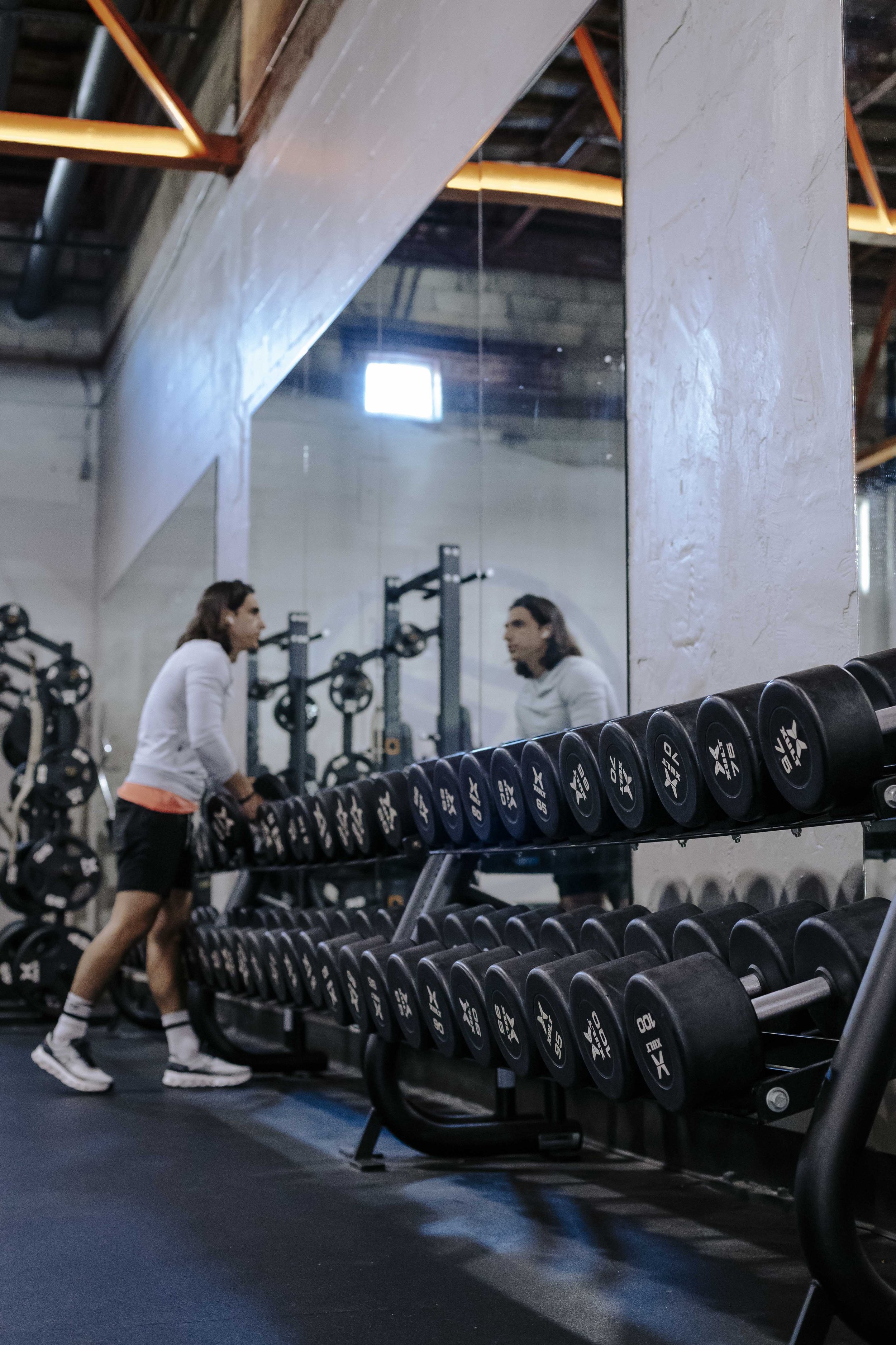 A woman is walking past a dumbbell rack in a gym, with a mirror reflecting her and the gym equipment.