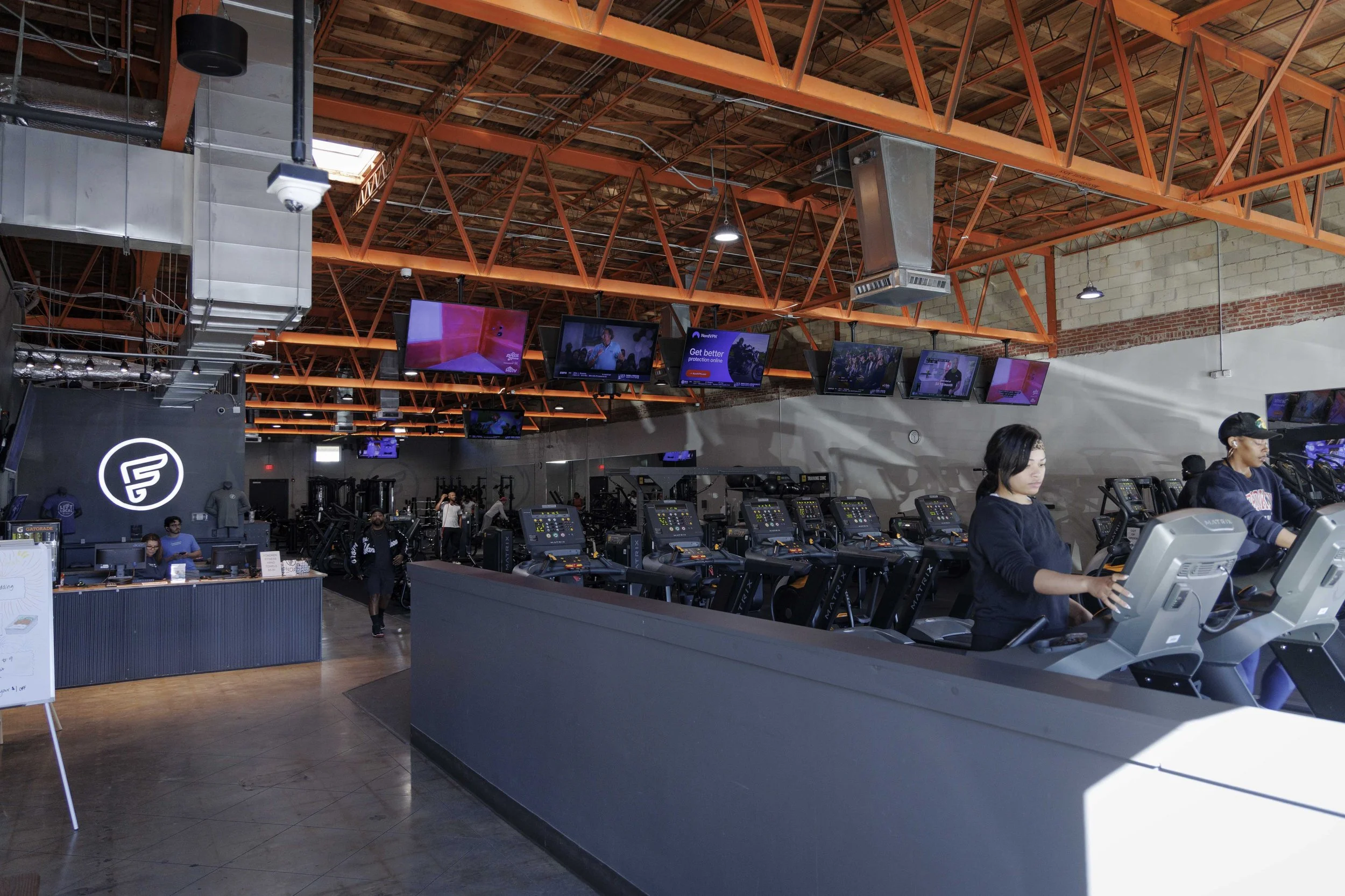 Inside a gym with high wooden ceiling and orange metal beams, several people are working out on treadmills, while a few others are at the reception counter. Multiple TV screens are mounted on the wall, displaying various programs.