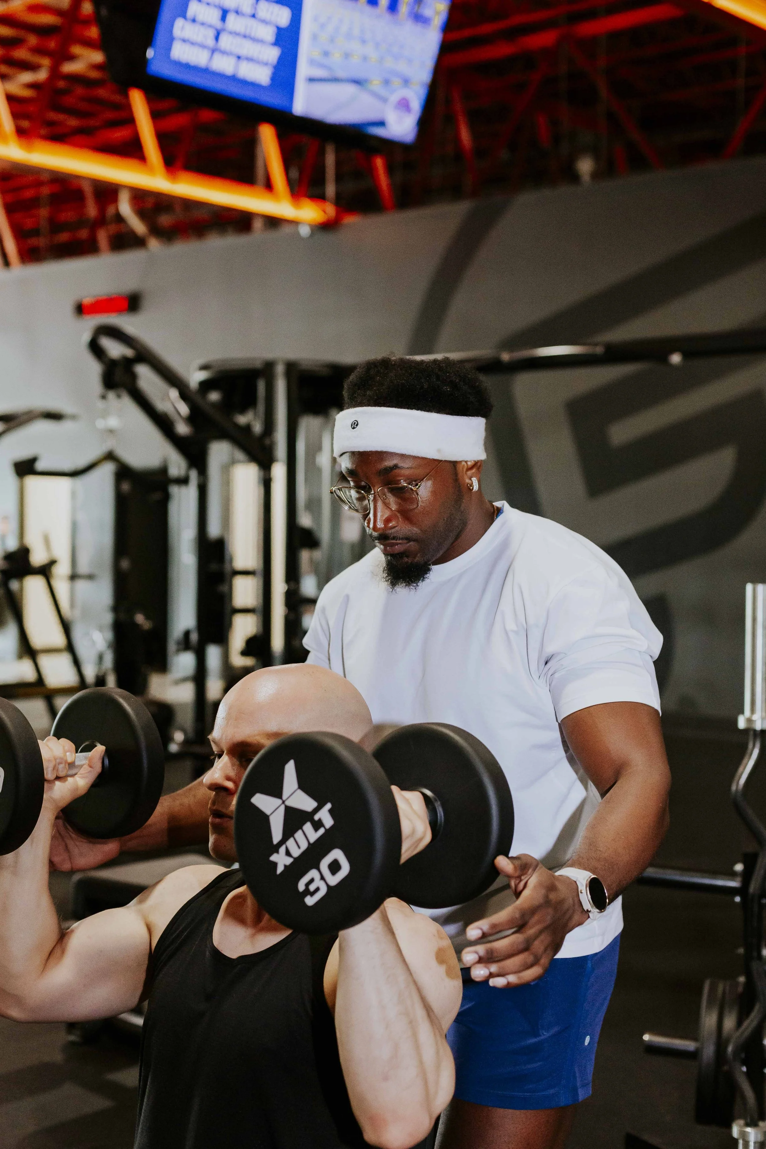 A personal trainer assisting a person doing a bicep curl with dumbbells at the gym.