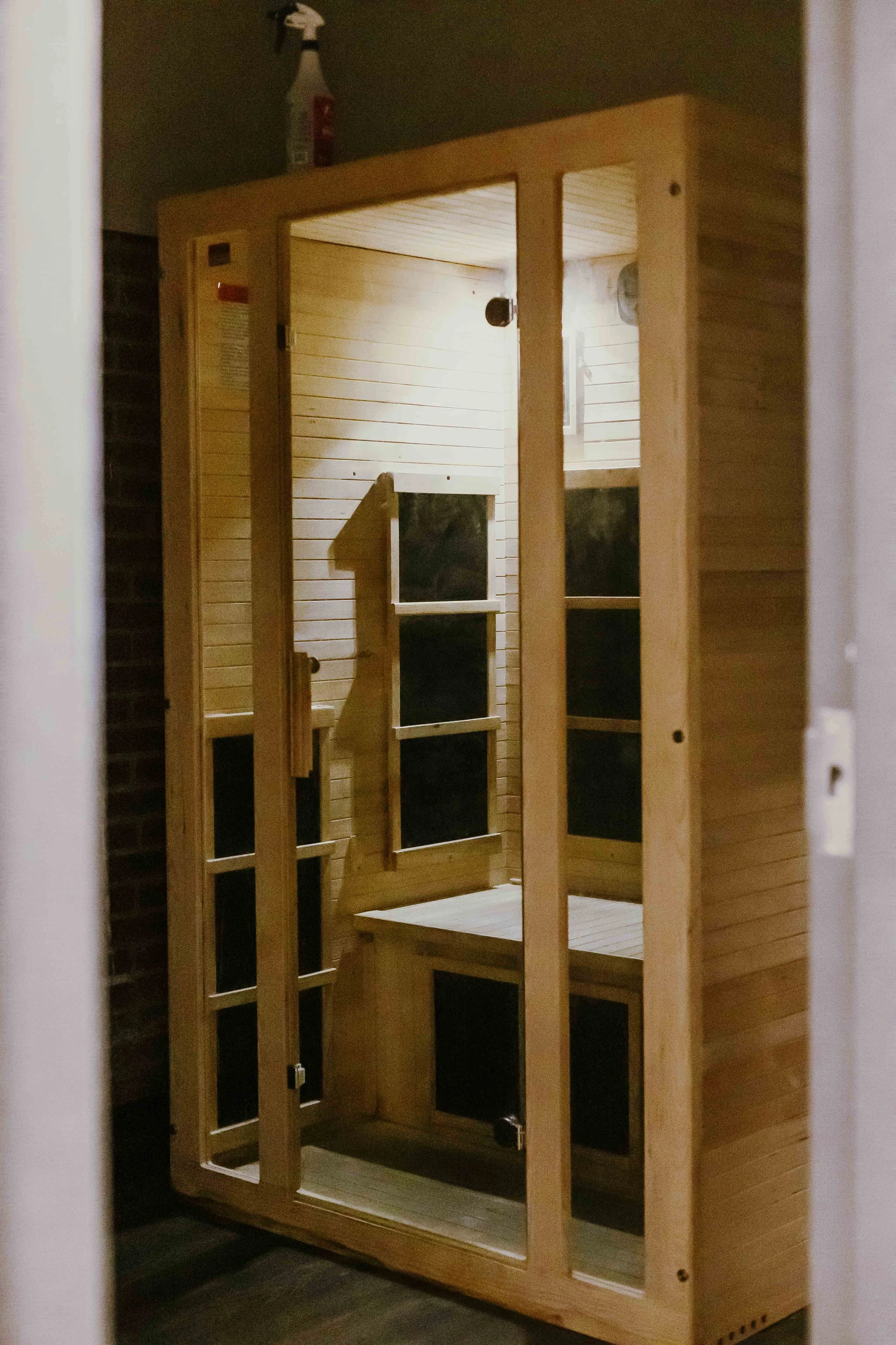 Unfinished wooden sauna in a room with dark walls. The sauna has a glass door, wooden benches, and lighting inside.