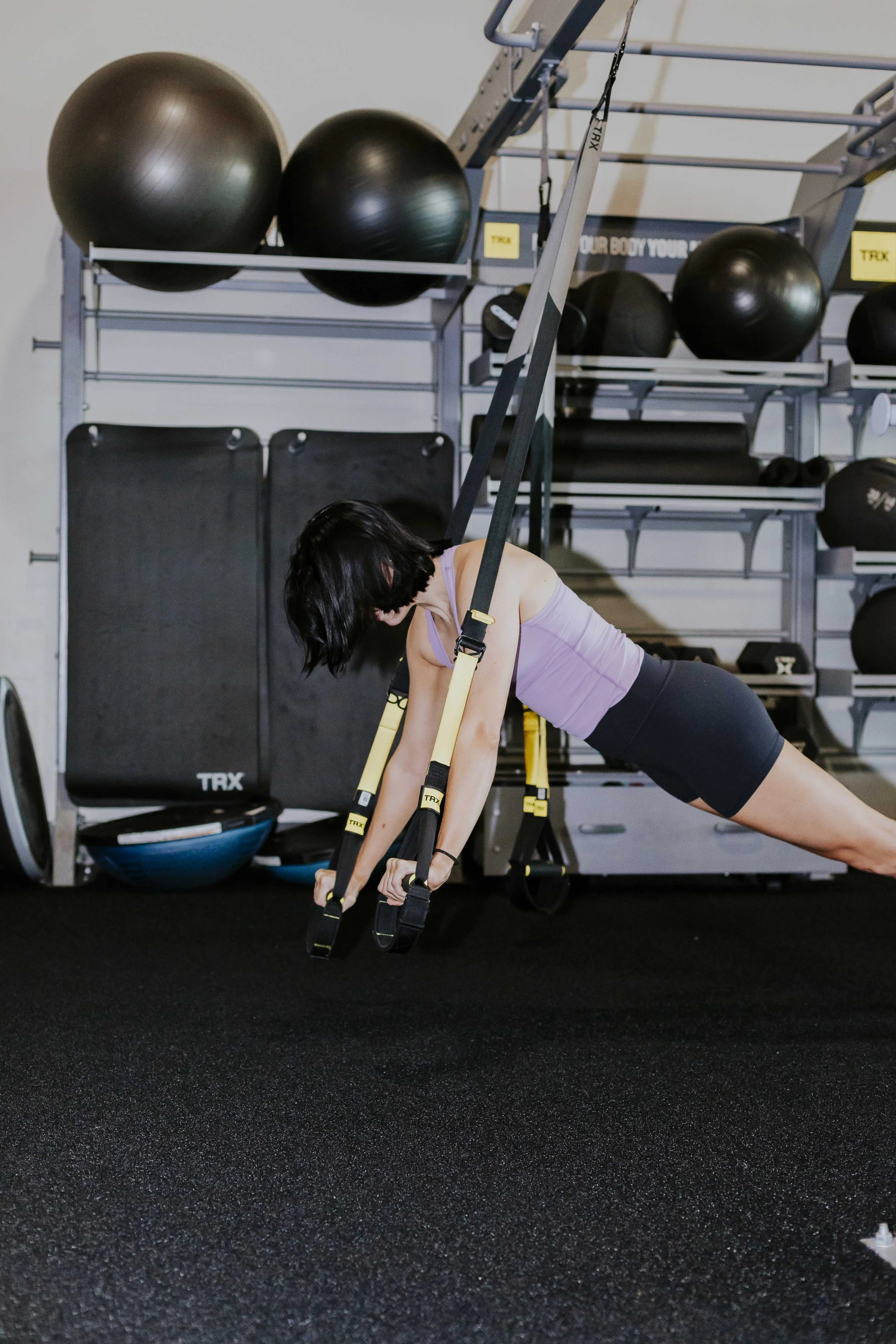 A woman in workout clothes is doing an incline plank exercise on a black gym mat using suspension straps in a gym. There are large exercise balls and workout equipment on shelves in the background.