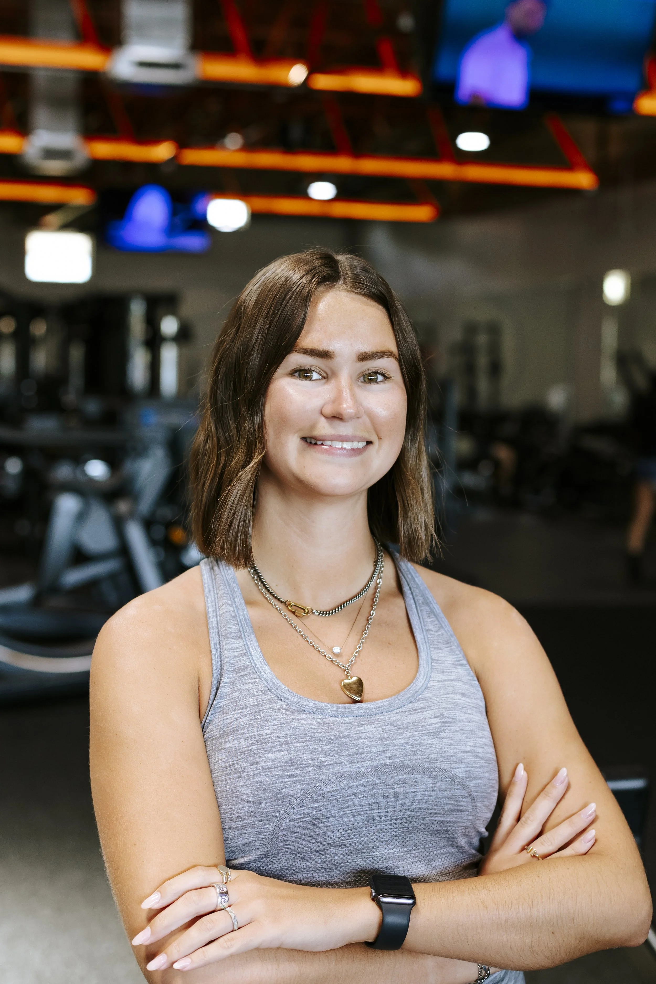 A young woman with shoulder-length brown hair, wearing a gray tank top, multiple necklaces, a smartwatch, and rings, standing with her arms crossed in a gym with exercise equipment in the background.