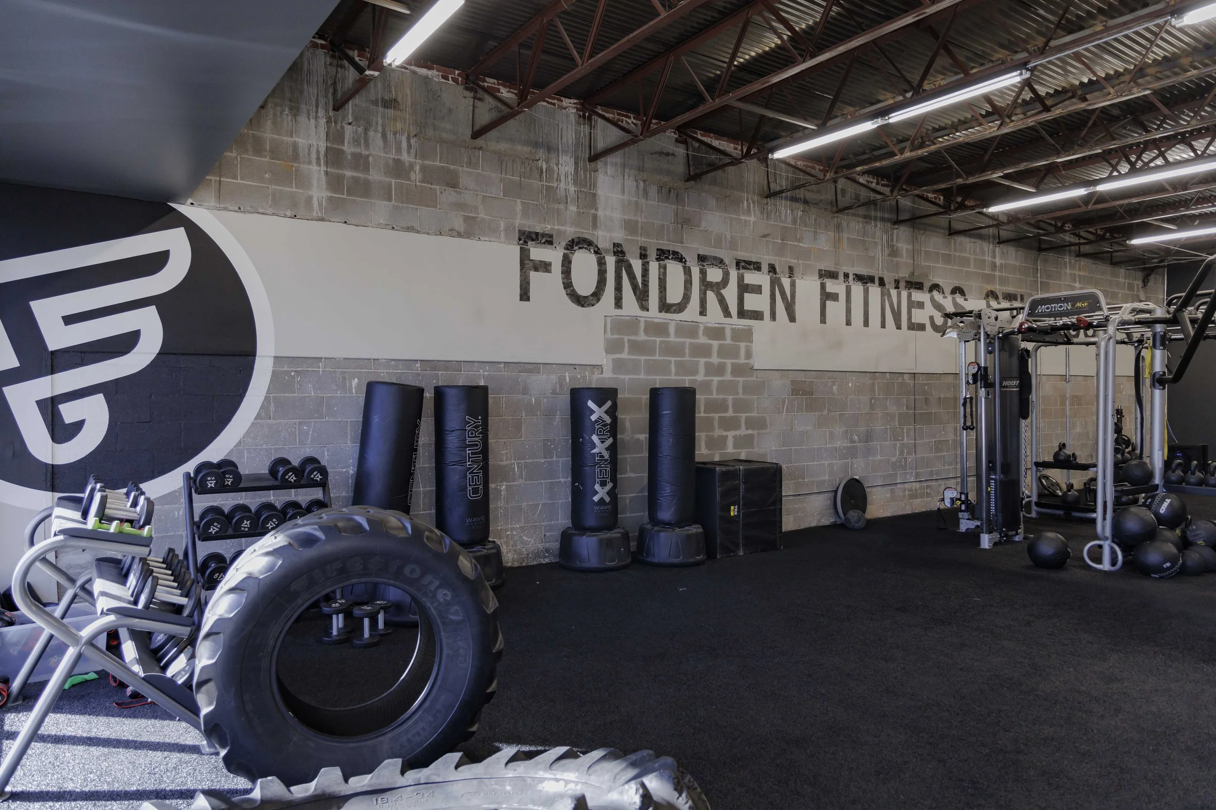 A fitness gym with weight training equipment, punching bags, and large tires in front. The wall has a logo and the words "FONDREN FITNESS" painted on it. There are dumbbells, medicine balls, and other gym gear visible.