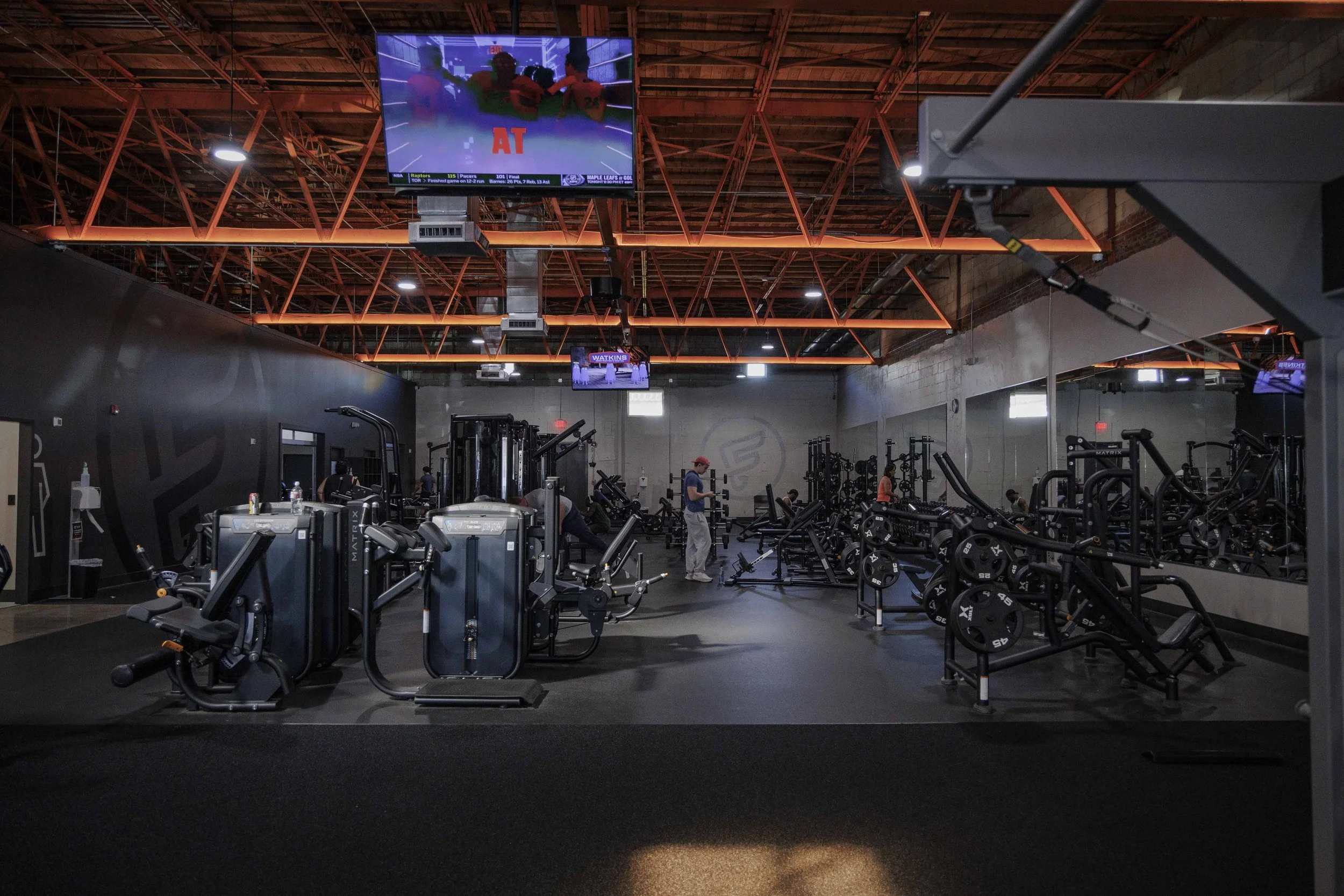 Interior of a gym with various exercise equipment and a few people working out, illuminated by orange lighting on the ceiling.