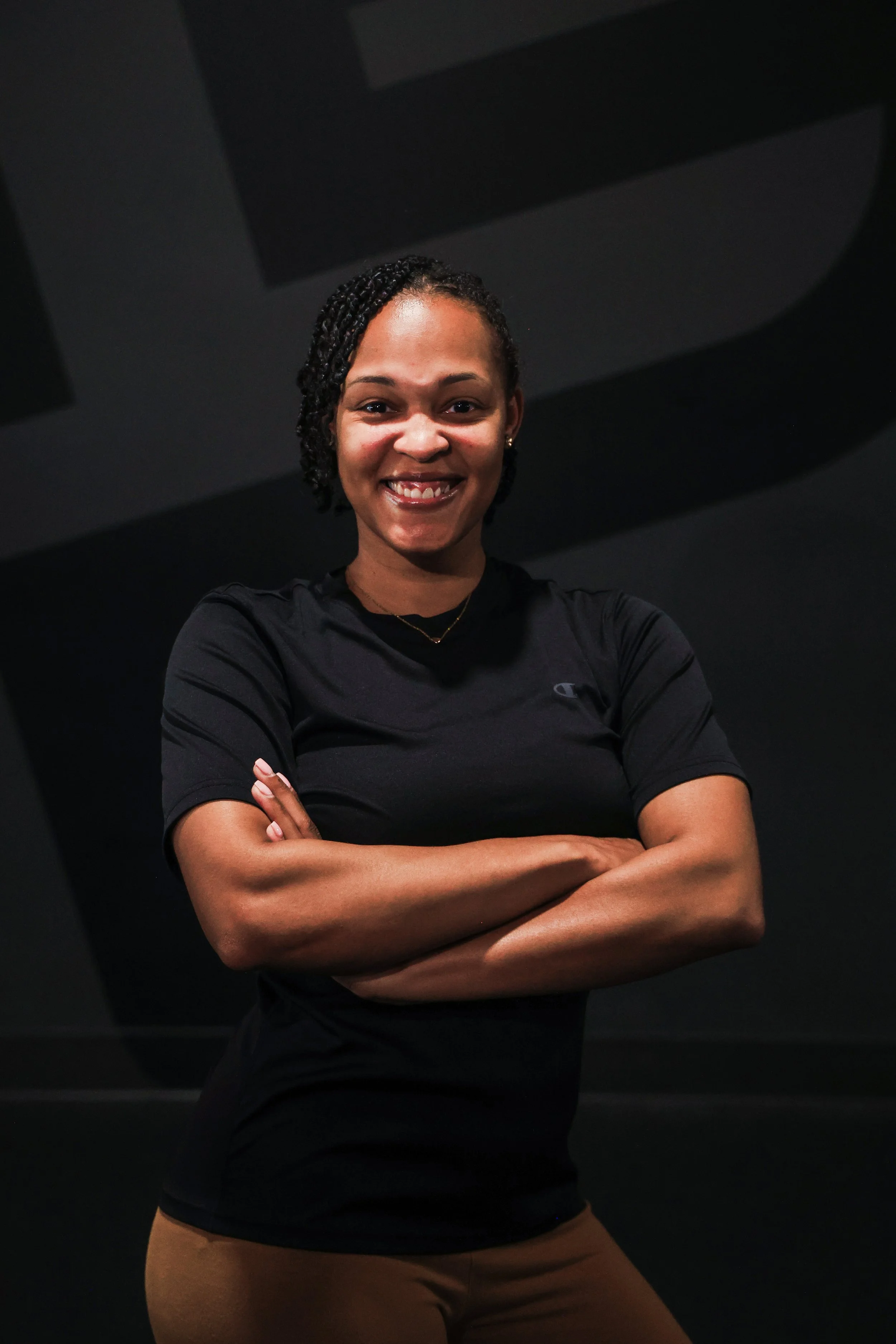 A young woman with curly hair styled in an afro and wearing a black zip-up jacket, standing with arms crossed, smiling in front of a large black wall with a geometric design.