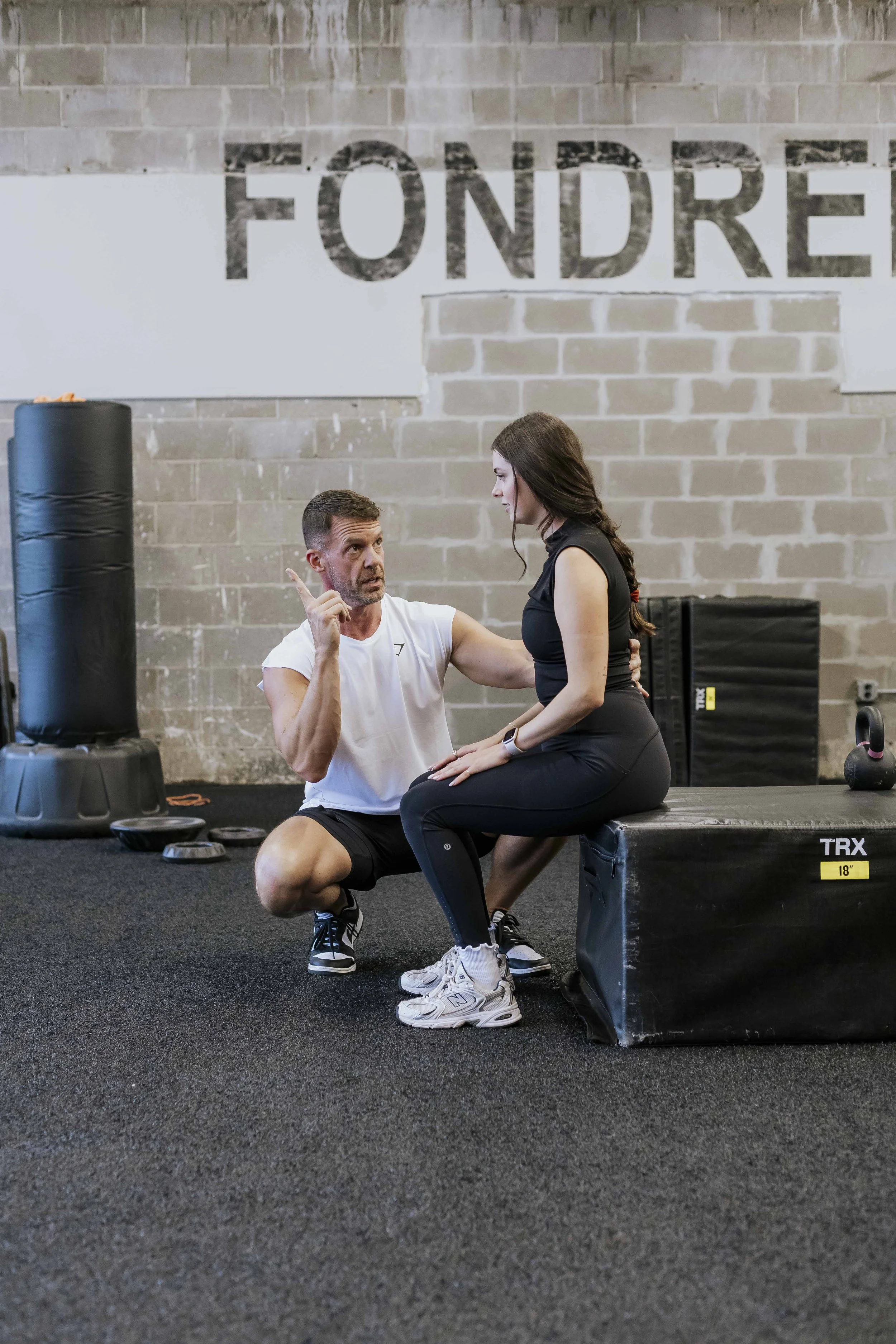 A trainer instructs a woman during a gym workout in a fitness facility with a brick wall background. The woman is seated on a black plyometric box labeled 'TRX 18"', wearing black workout clothes and white sneakers, while the trainer, dressed in a white T-shirt and black shorts, is kneeling next to her, emphasizing a point.