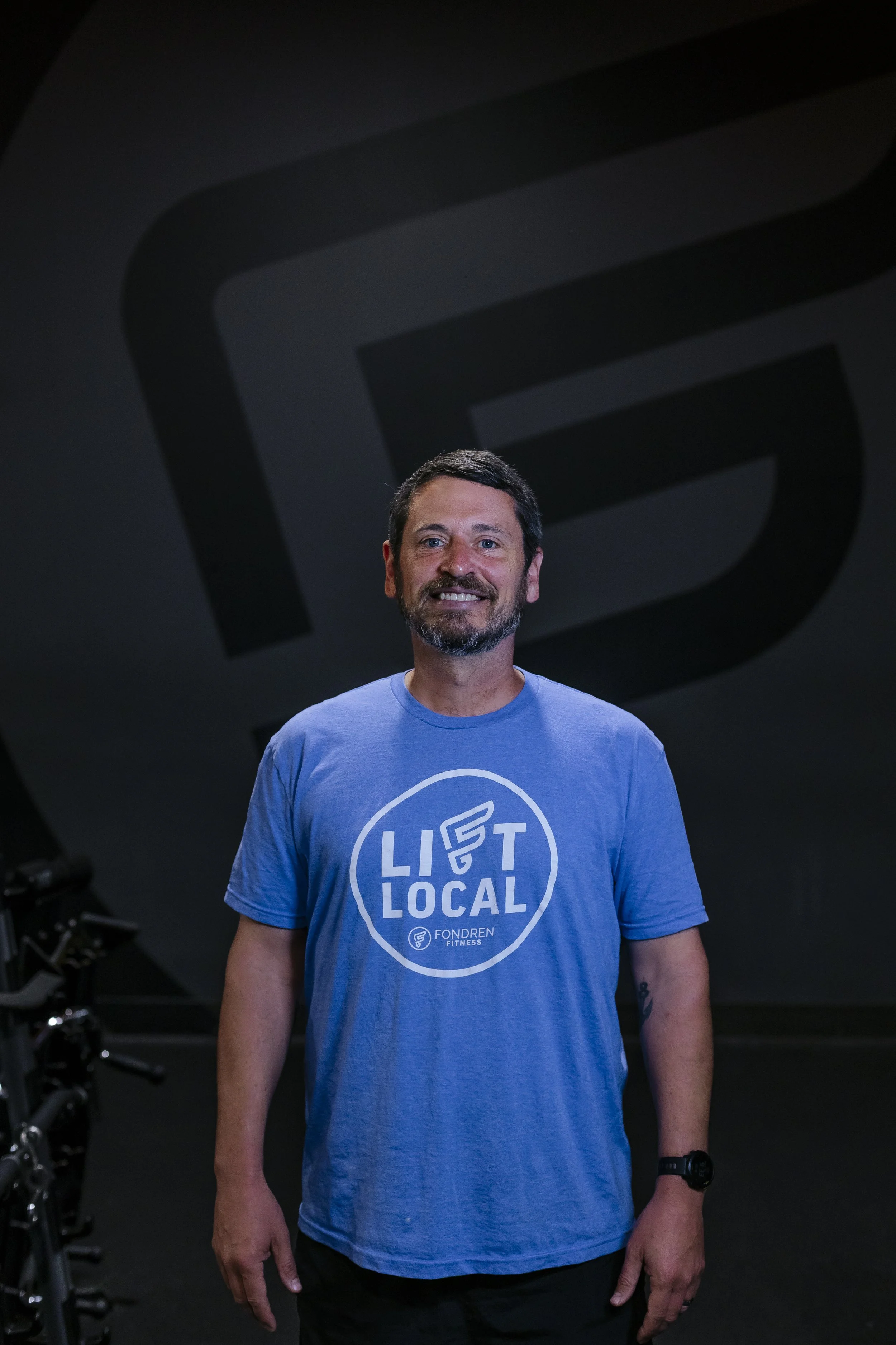 A man with a beard and short dark hair smiling, wearing a light blue t-shirt with a circular logo that reads 'LIFT LOCAL' and 'FONDREN FITNESS,' standing in a dark gym with fitness equipment and a large black wall in the background.