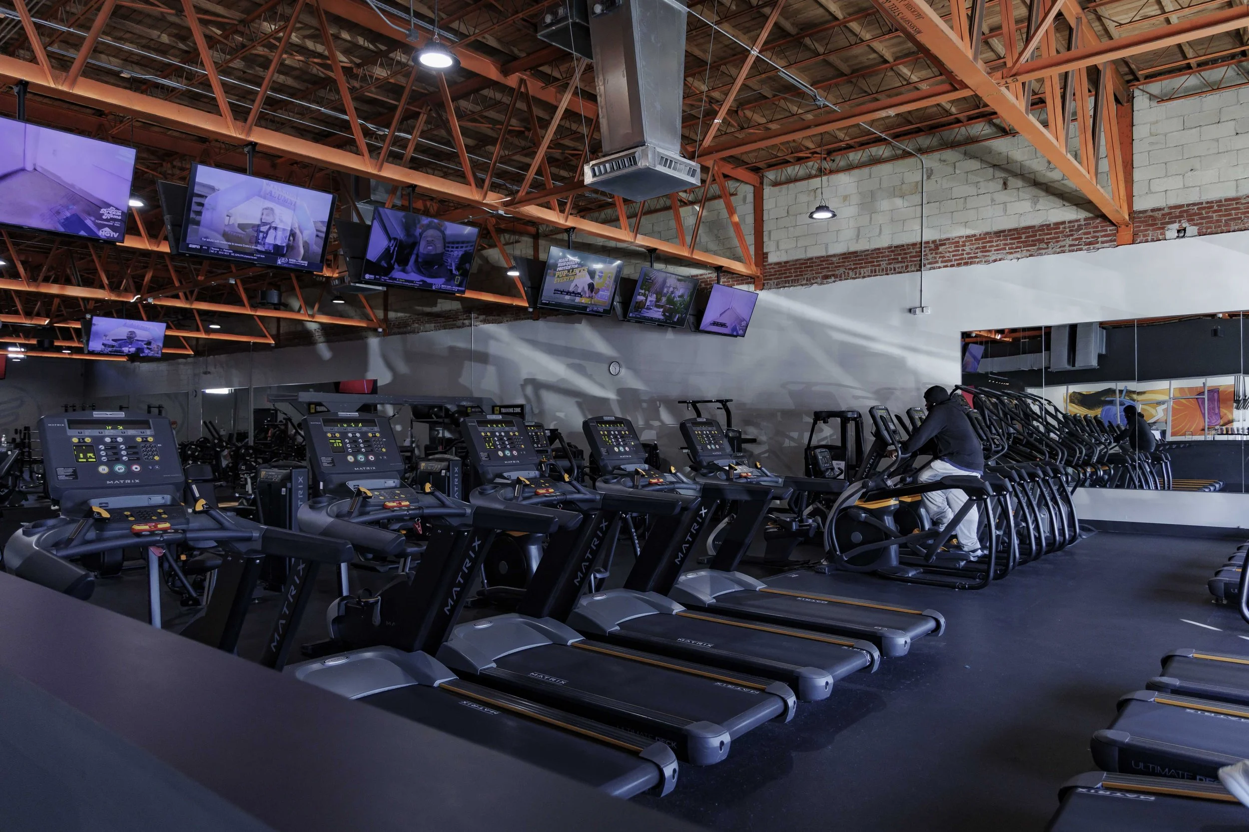 Indoor gym with multiple treadmills and wall-mounted TVs, with exposed brick and metal beams ceiling.