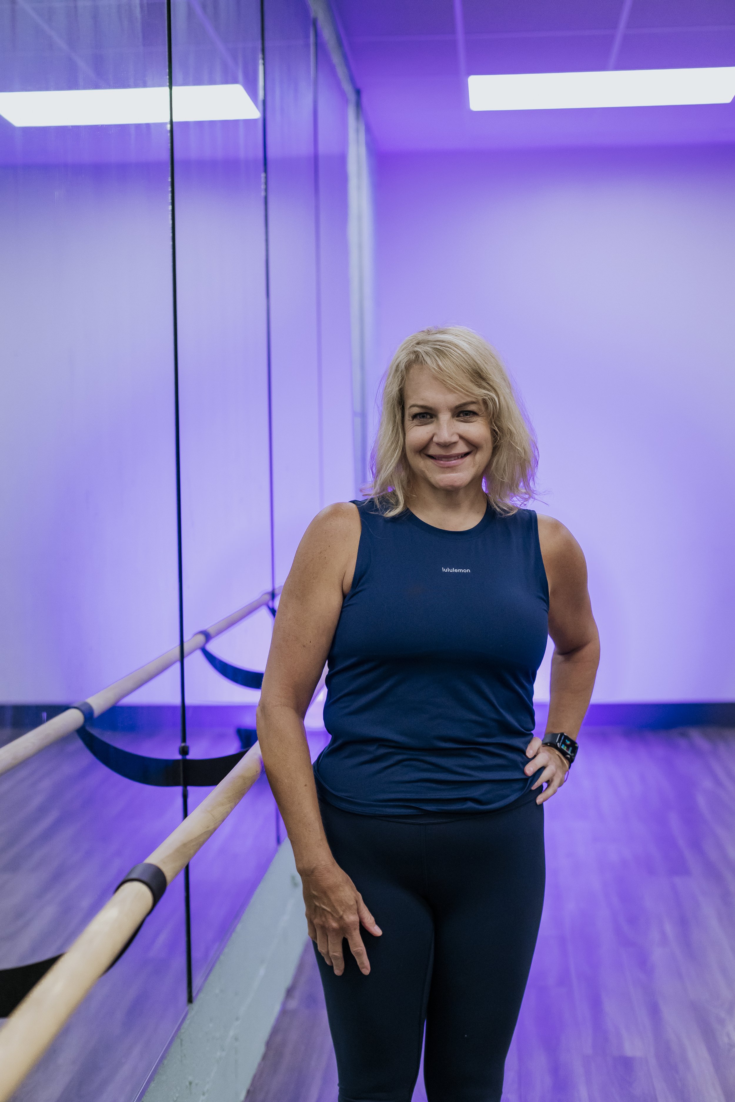 A woman with blonde hair wearing a navy blue sleeveless top and matching pants, smiling and posing in a dance studio with purple lighting and mirrored walls.