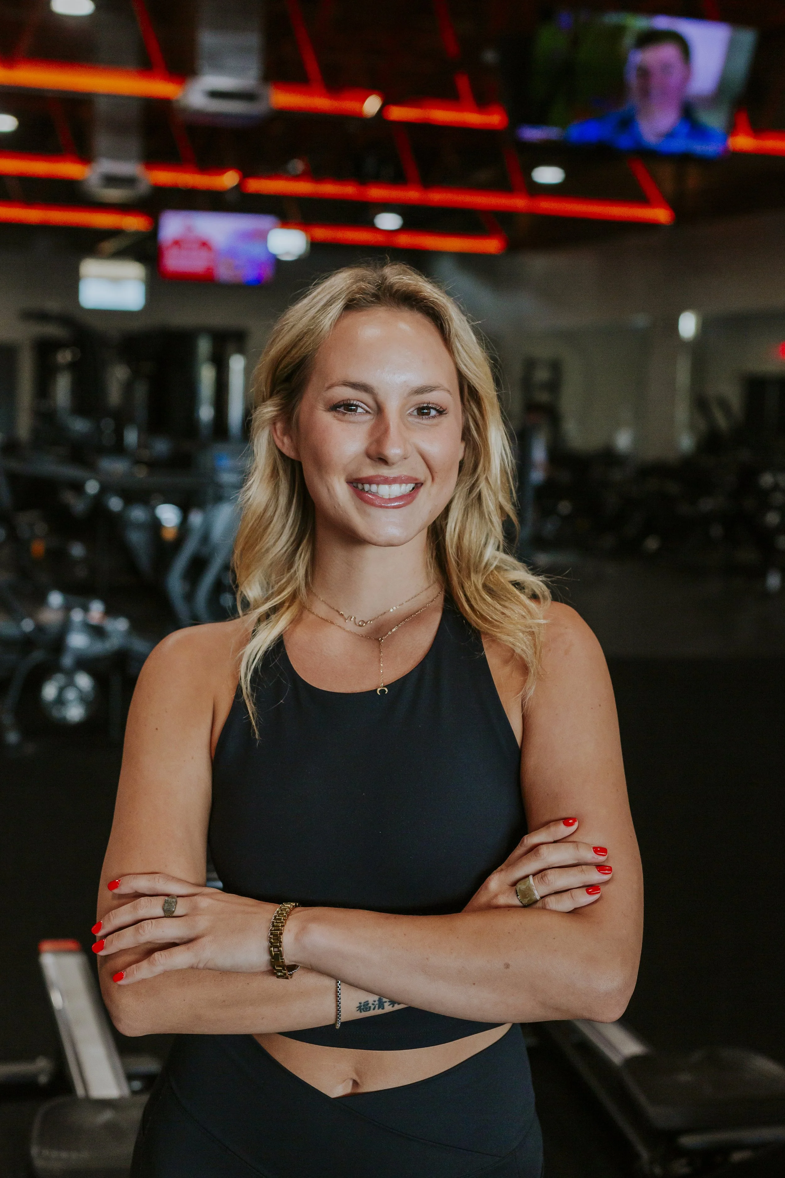 A young woman with blonde hair smiling with arms crossed in a gym setting, wearing a black sports top and jewelry, with gym equipment and TV screens visible in the background.