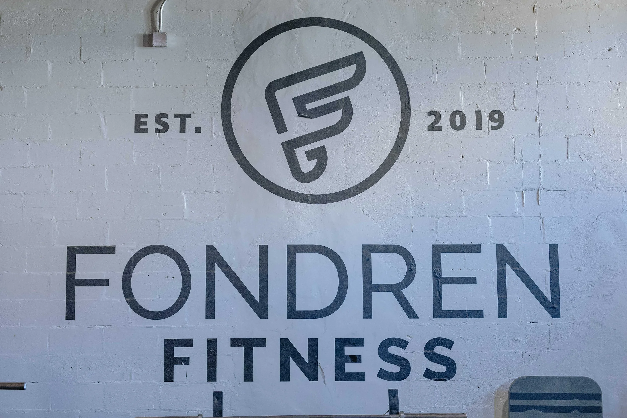 A wall with a logo that resembles a stylized letter 'F' inside a circle, with the words 'EST. 2019' to the left and right of the logo. There is also part of the word 'FOUNDER' visible at the bottom and the word 'FITNESS' below it.