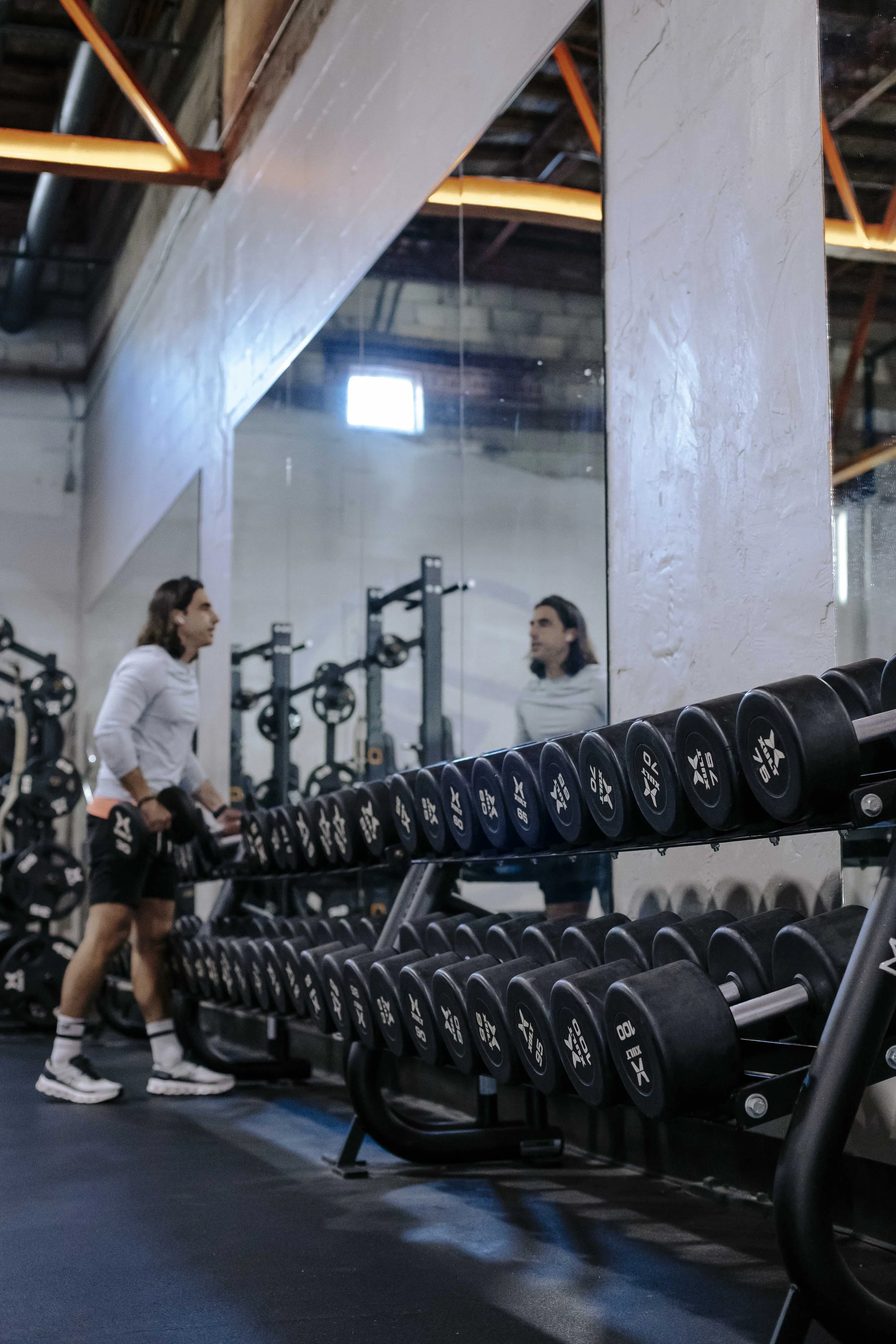 A woman with long hair wearing a white shirt and black shorts standing in a gym, facing a mirror, with dumbbells stored on a rack in front. The gym has a rustic ceiling with exposed beams and a large mirror reflecting the woman and some gym equipment
