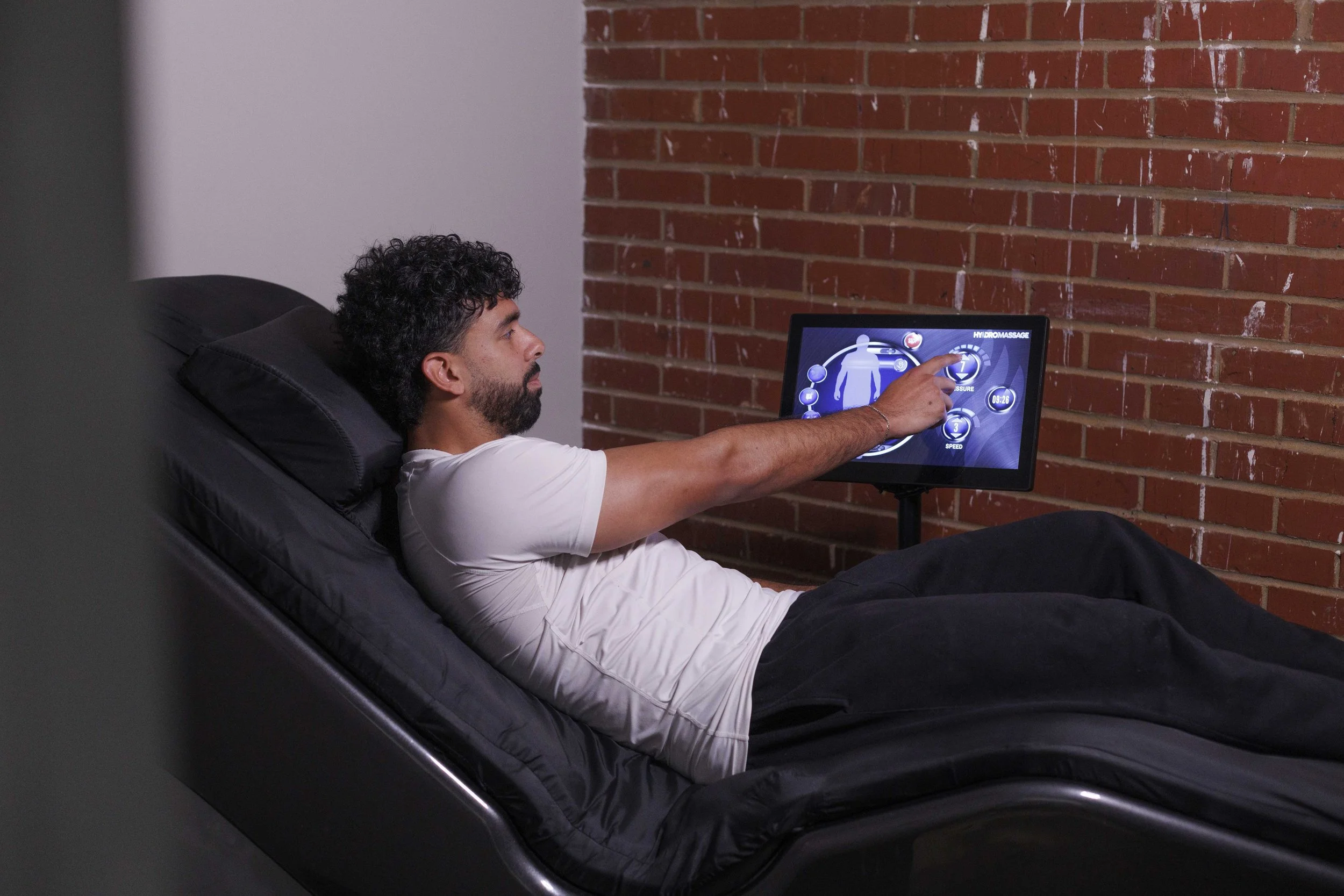 A man with curly dark hair and a beard is sitting on a black recliner chair, reaching out to a touchscreen device that displays health data, with a red brick wall behind him.