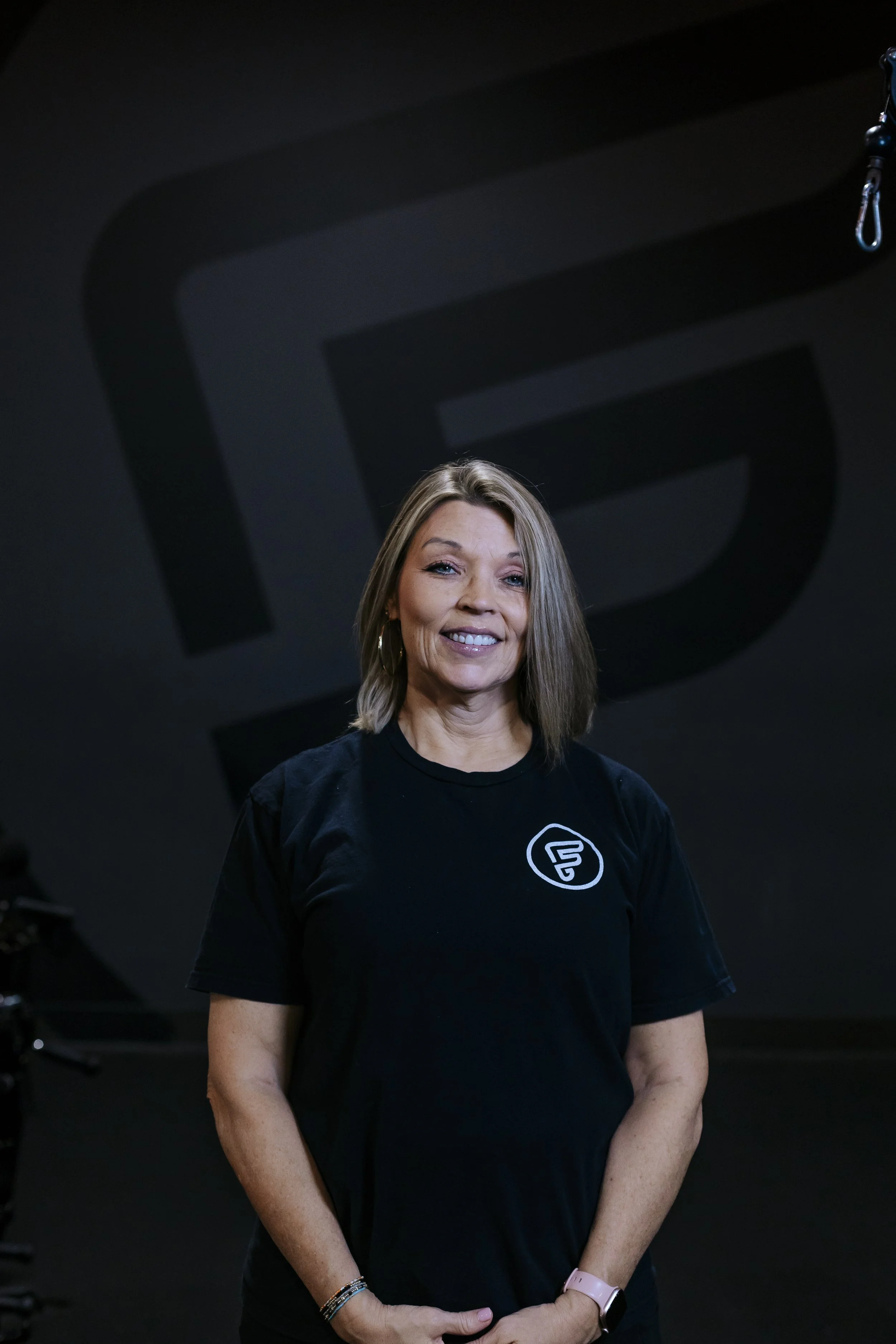 A woman standing in a dark gym, smiling, wearing a black T-shirt with a logo, with her arms crossed in front of her.