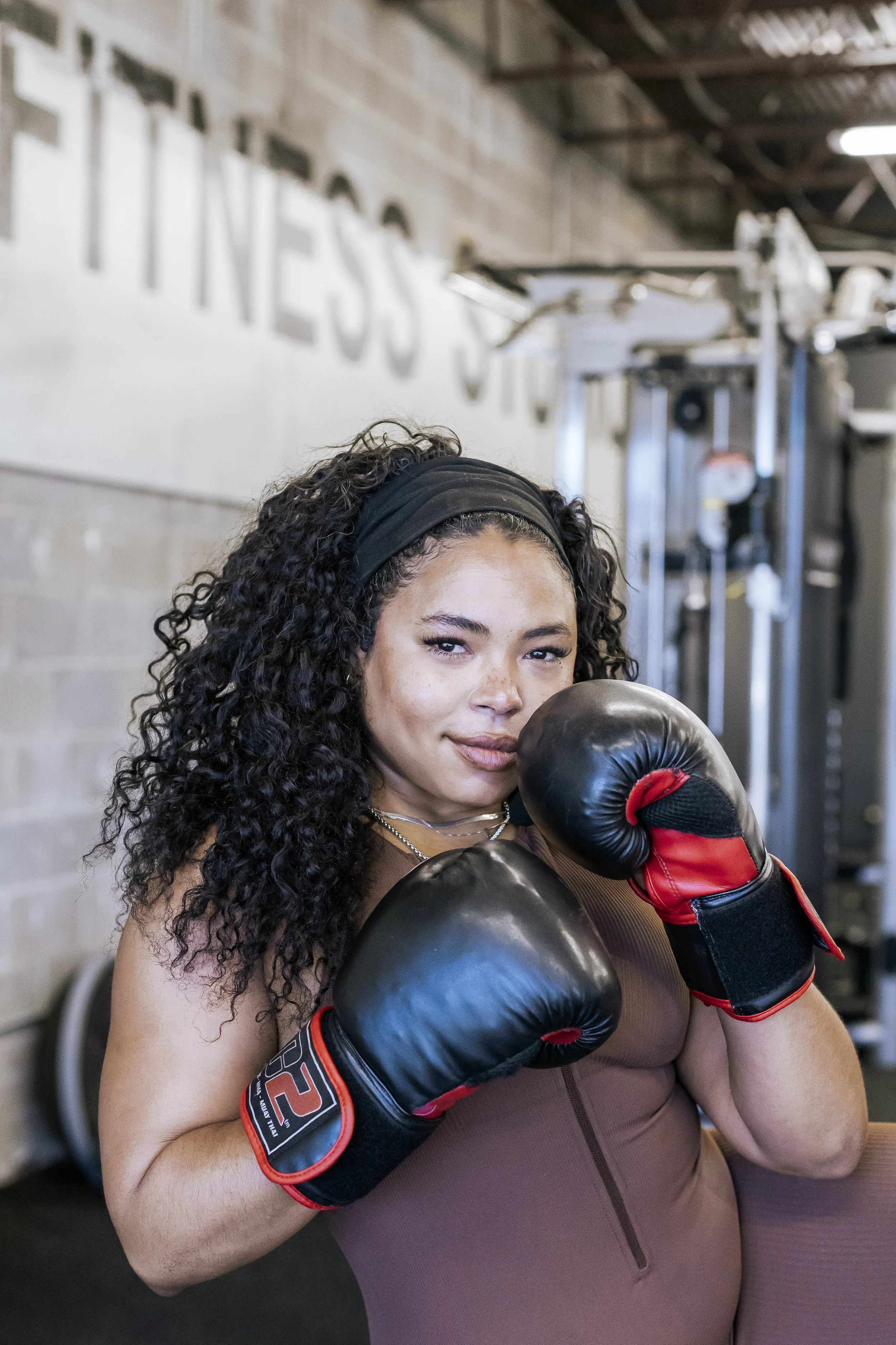 A woman with curly hair wearing gym gloves and workout clothing, standing in a gym, with her arm raised in a boxing stance.
