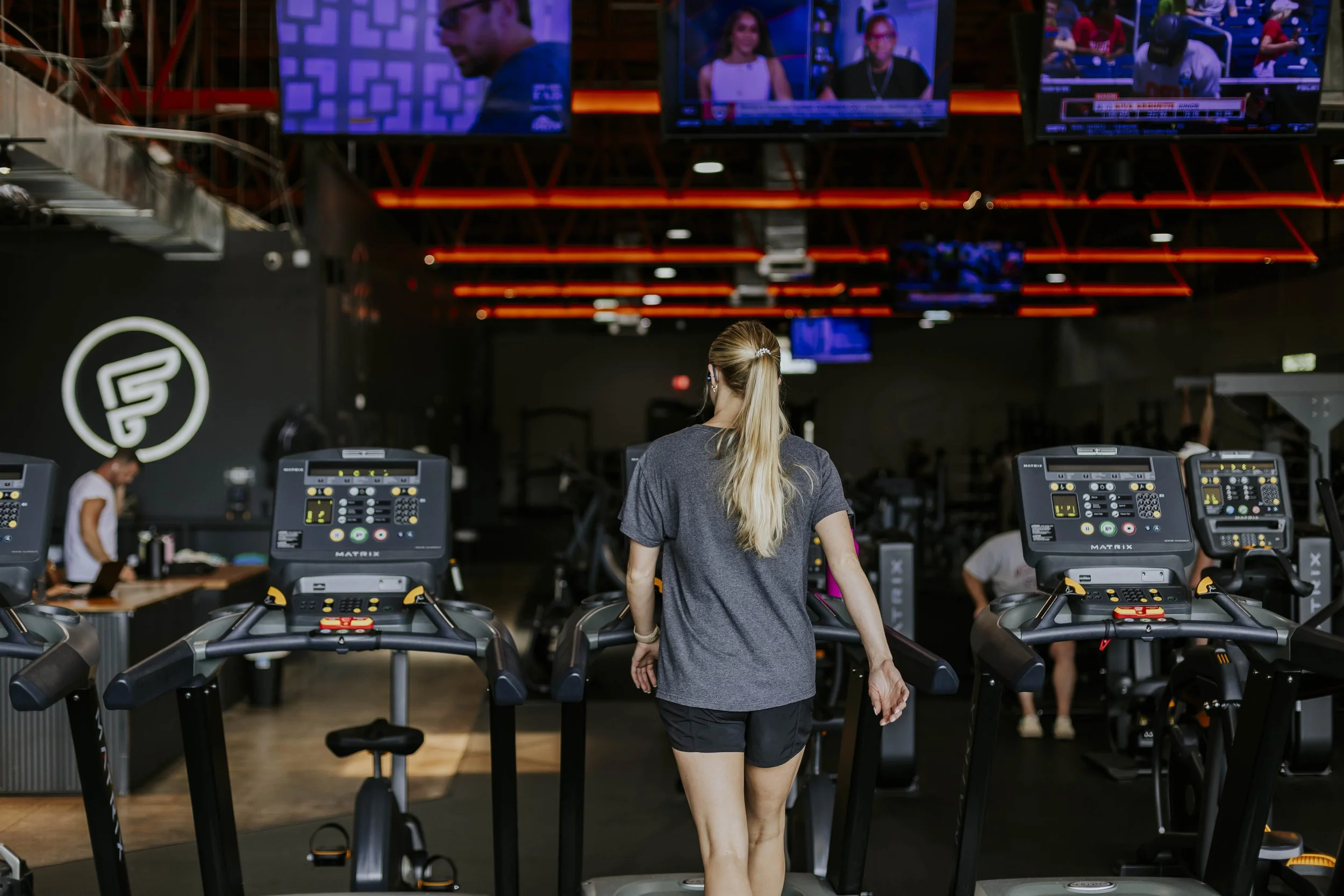 A woman with long blonde hair wearing a gray shirt and black shorts walks on a treadmill in a gym, surrounded by exercise equipment and large display screens.