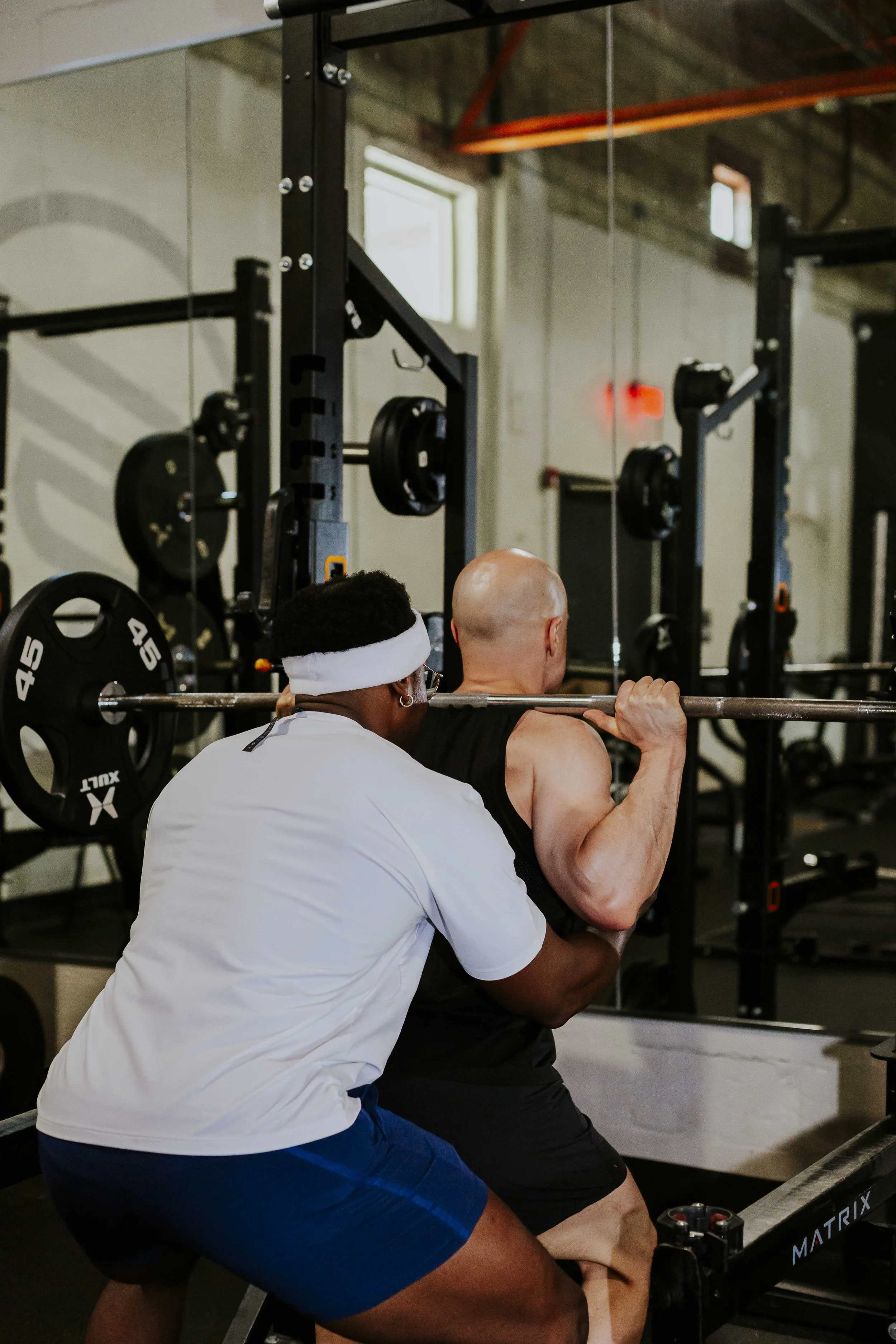 A personal trainer assisting a man with a barbell squat exercise in a gym, with weightlifting equipment visible in the background.