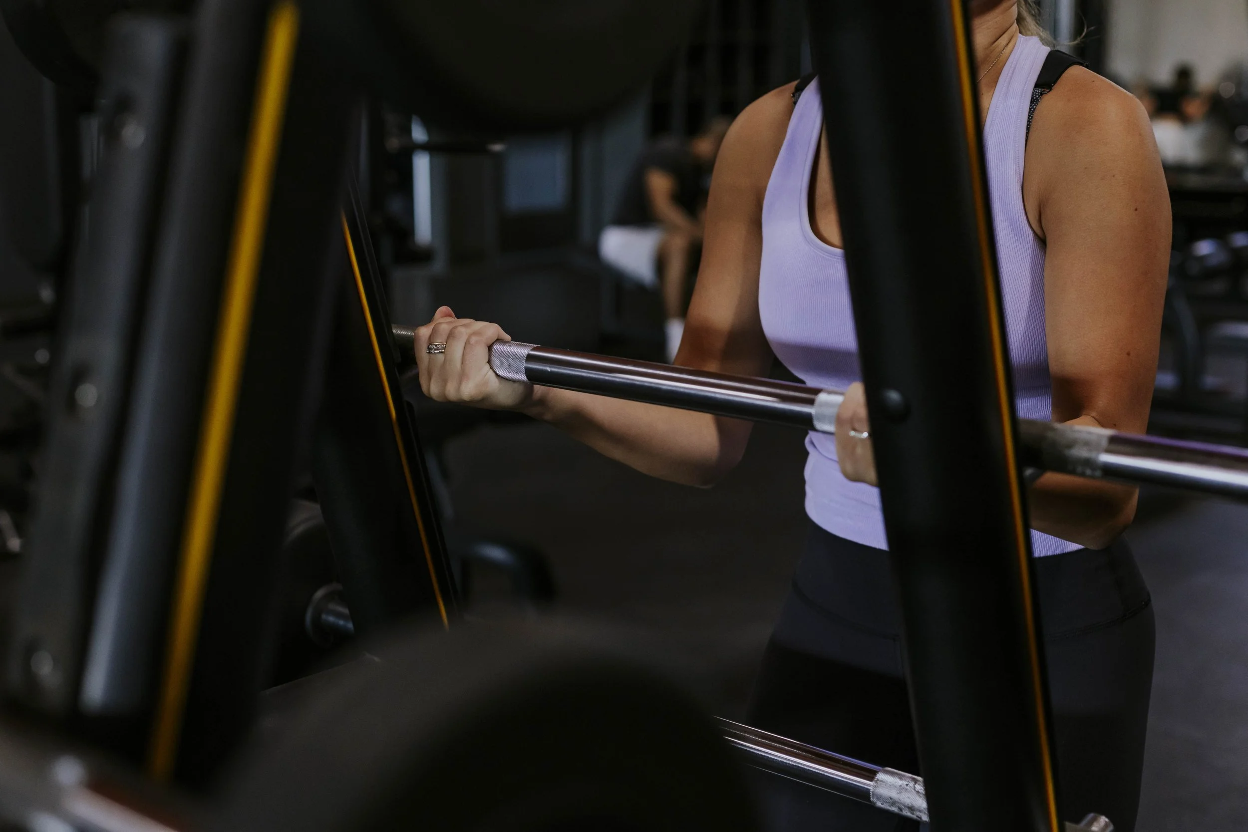 A person lifting a dumbbell at the gym, with focus on their arm and hand, wearing a white sleeveless top.