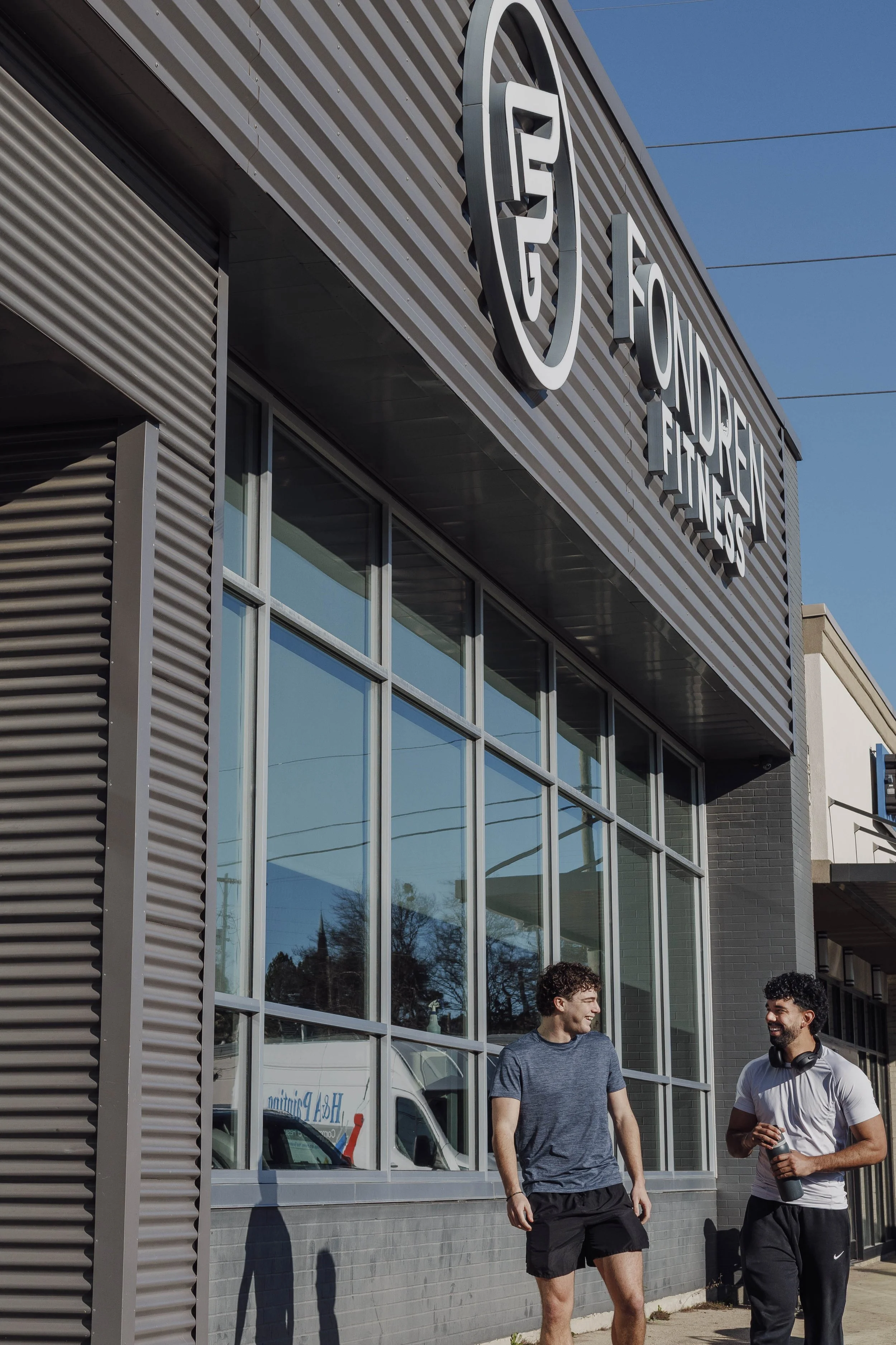 Two young men talking outside a modern building with large glass windows and a horizontal metal siding facade. The sky is clear and blue. One man has headphones around his neck, and a white van is partially visible inside the building.