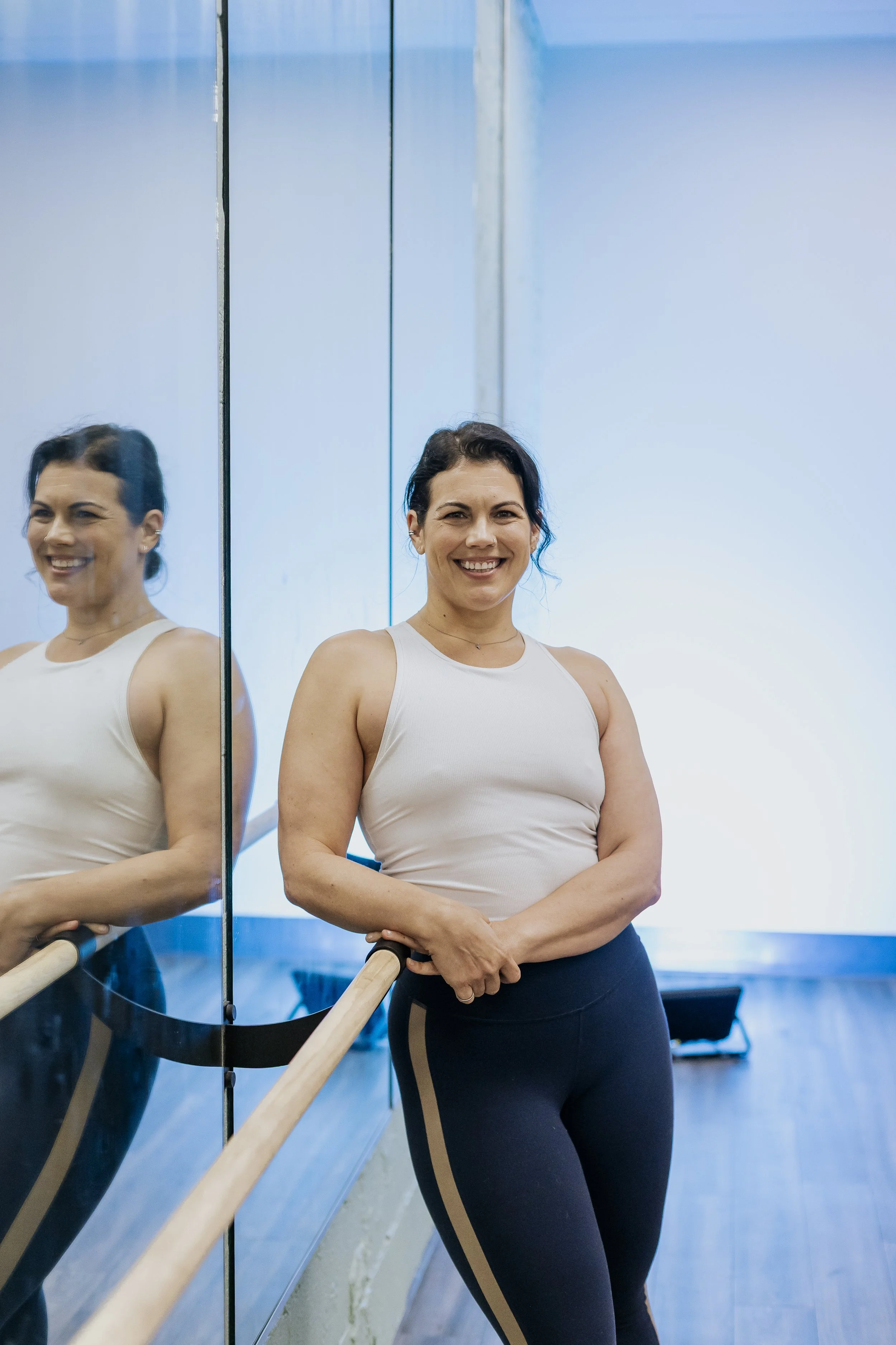 A woman standing at a ballet barre in a dance studio, smiling, with her reflection visible in a mirror on the left side of the image.