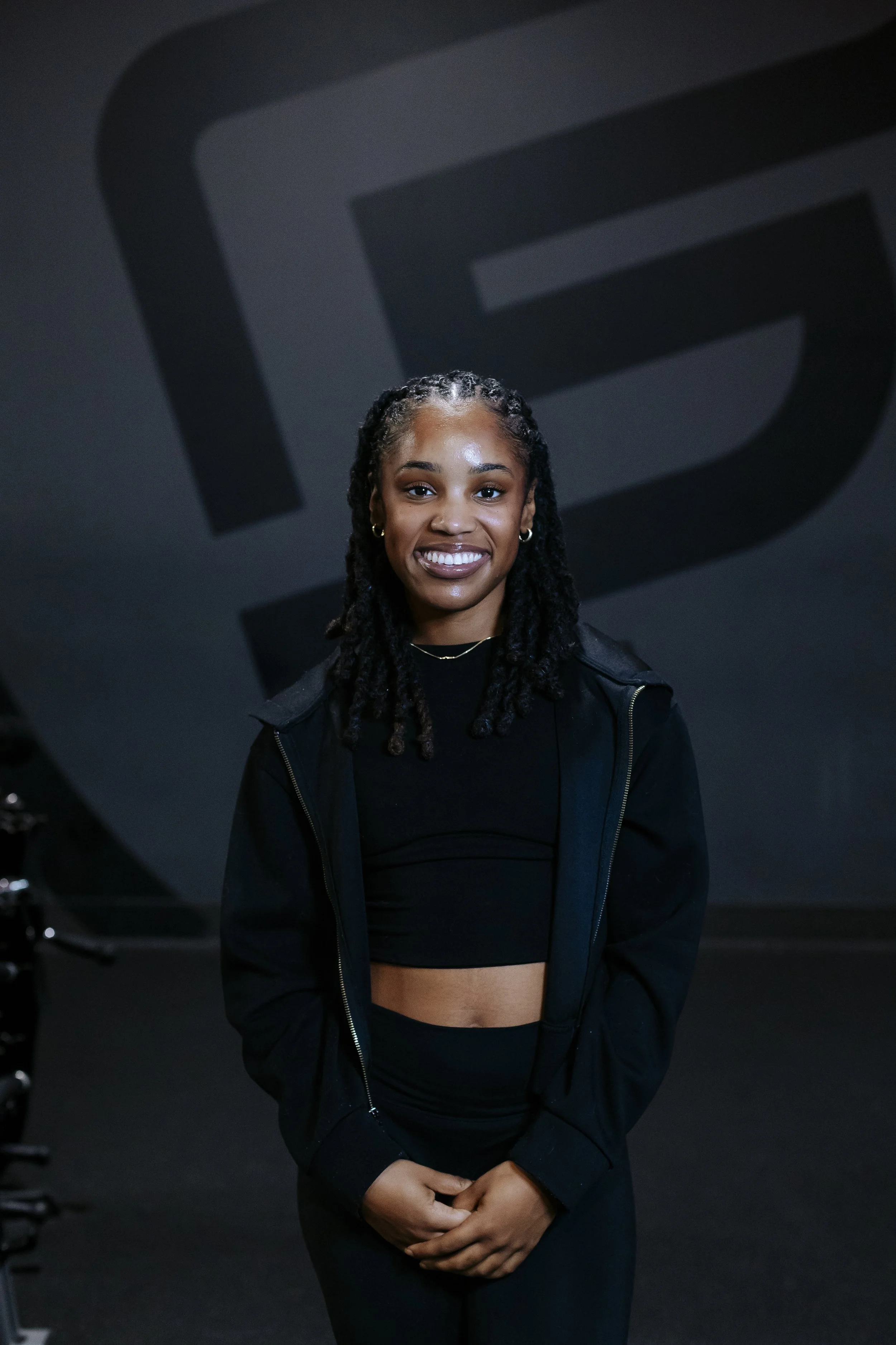 Young woman with braids wearing a black crop top, black jacket, and black pants, standing in front of a dark background with a large abstract black and gray logo.