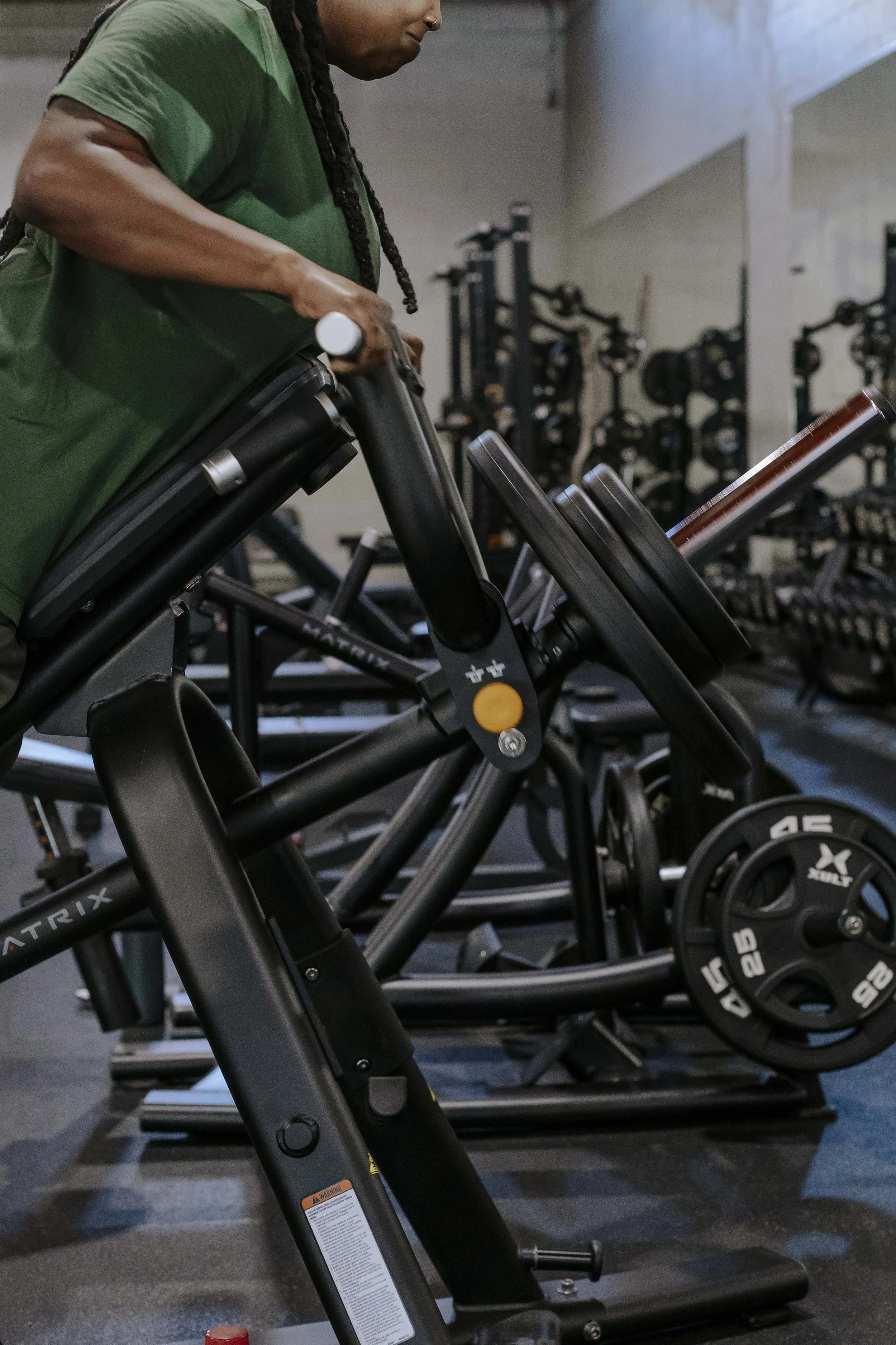 A person in a green shirt working out on a black elliptical trainer in a gym with multiple barbells and weight plates in the background.