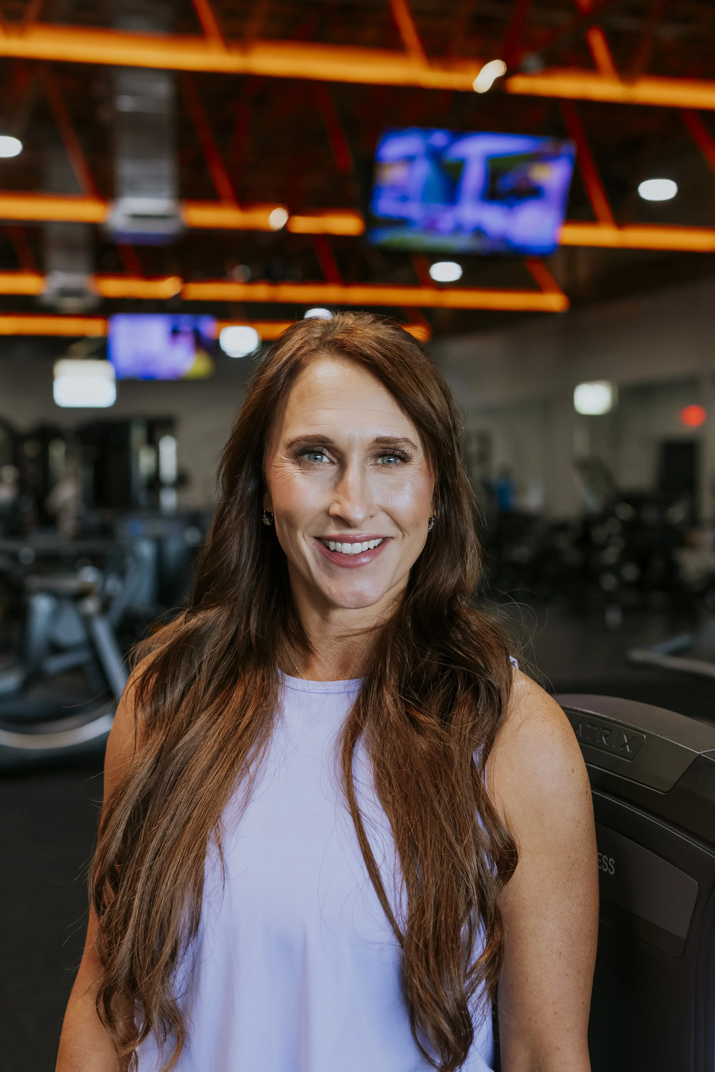 A woman with long brown hair and blue eyes smiling at the camera inside a gym, with exercise equipment and TV screens in the background.
