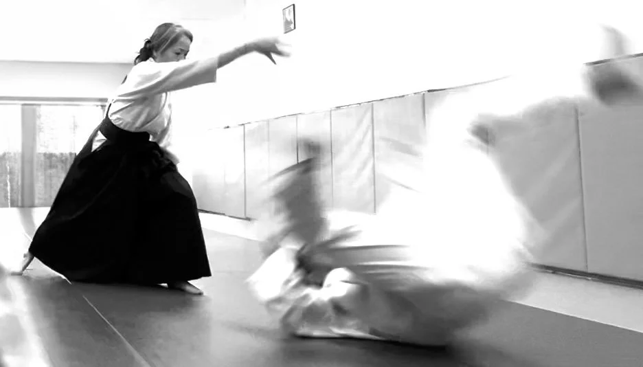 Two martial artists practicing judo in a dojo, one executing a throw while the other is falling to the mat, in black and white.