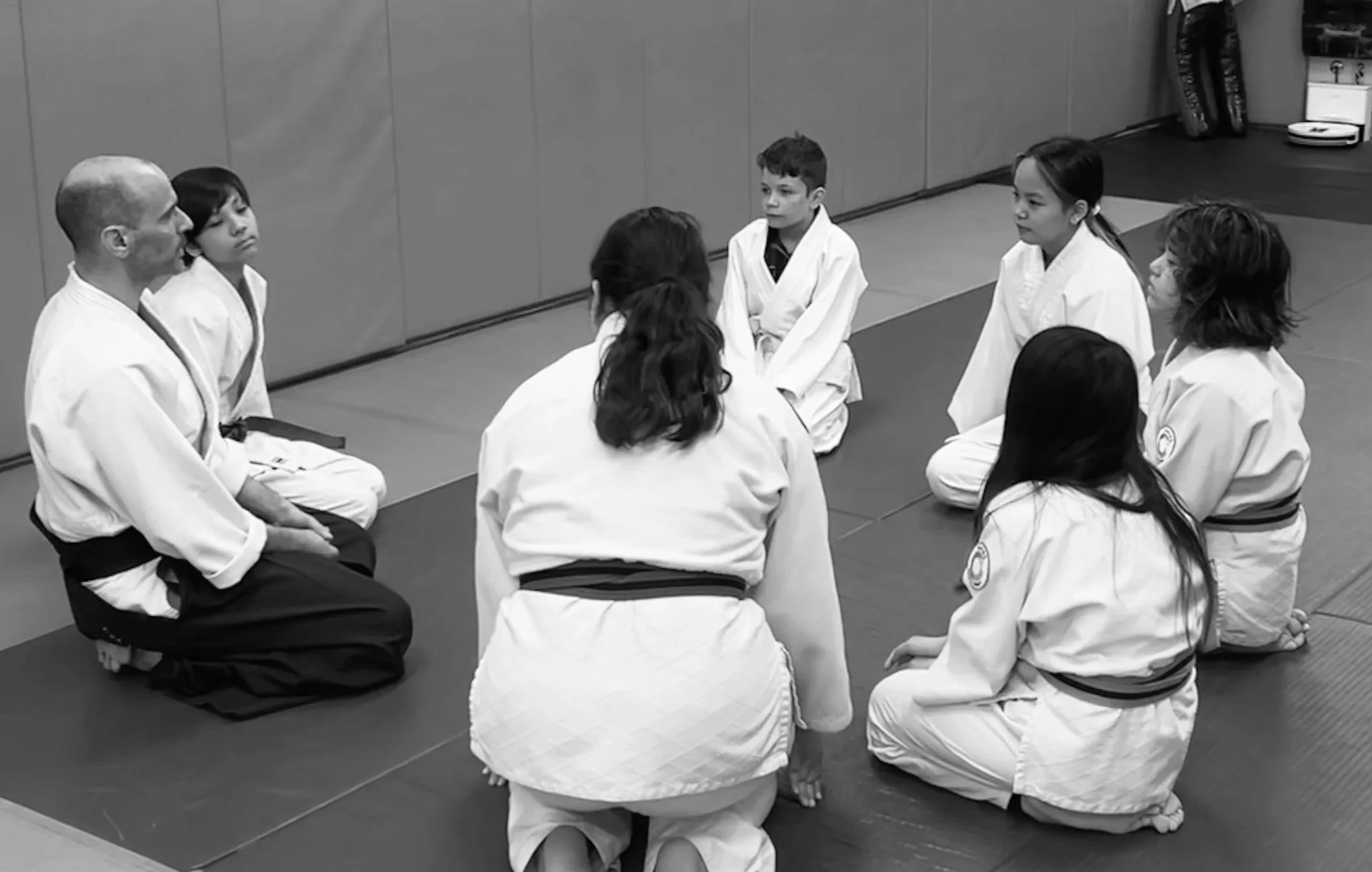 A group of children and their instructor, all dressed in white martial arts uniforms, seated in a circle on a gym mat during a martial arts class.
