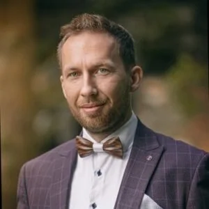 A man outdoors wearing a suit and bowtie, with a blurred natural background.
