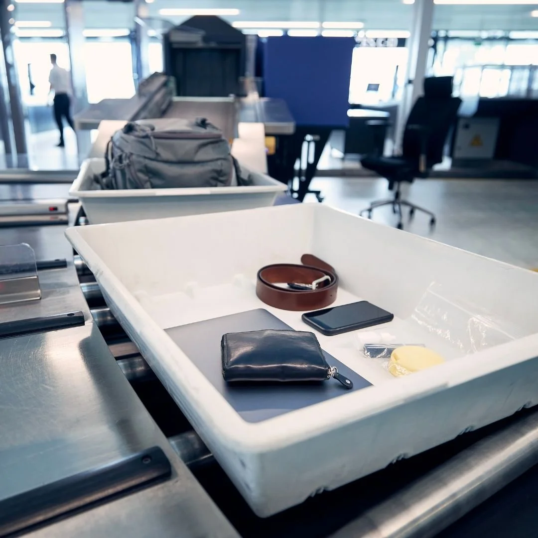 Items on a conveyor belt in an airport security checkpoint, including a black wallet, a brown belt, a smartphone, a bar of soap, and a small clear container, with a background of airport chairs and staff.