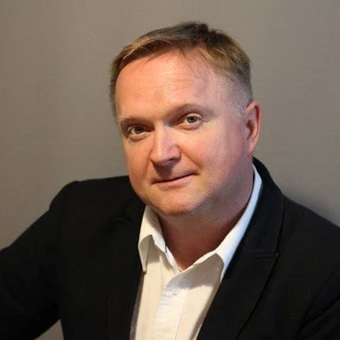 A man with short blond hair wearing a white shirt and black blazer, sitting against a gray background.