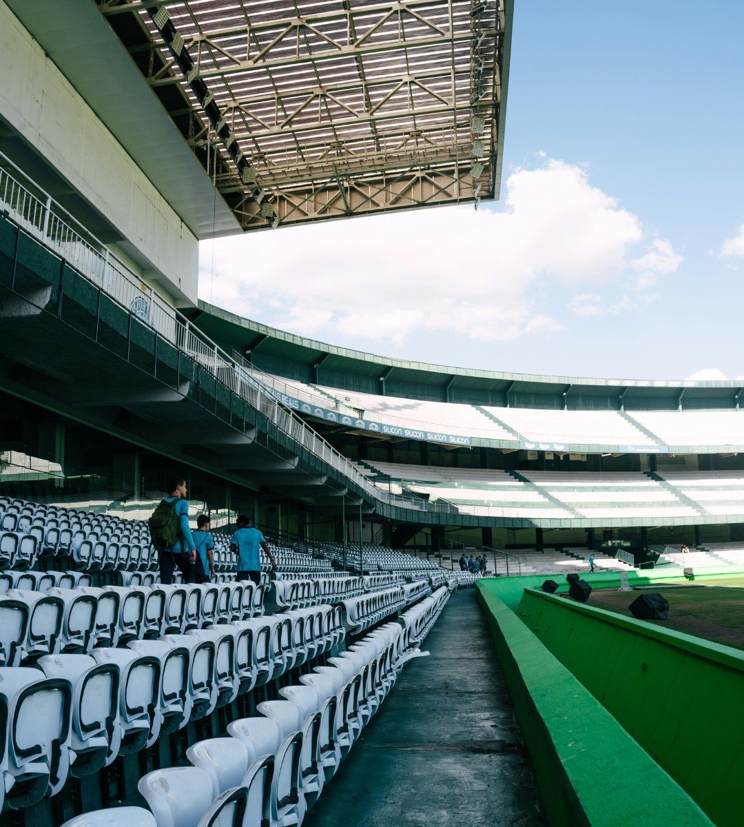 An empty baseball stadium with seating, a few people walking on the steps, and a green field under a blue sky with clouds.