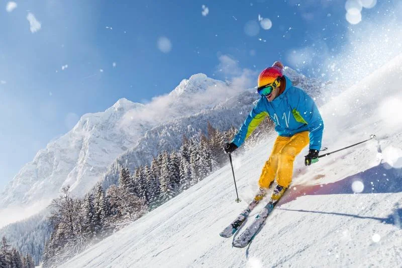 A person skiing down a snowy mountain slope with snow-covered trees and mountain peaks in the background, wearing a blue jacket, yellow pants, a rainbow-colored hat, and sunglasses.