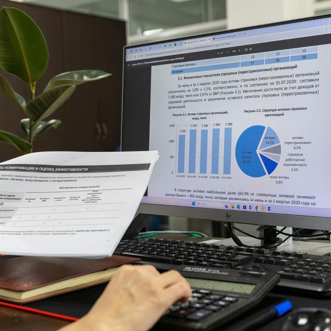 Computer monitor displaying financial data including bar charts and pie charts, a person's hand on a calculator, printed documents, a closed notebook, and a pen on a desk, with a plant and office environment in the background.